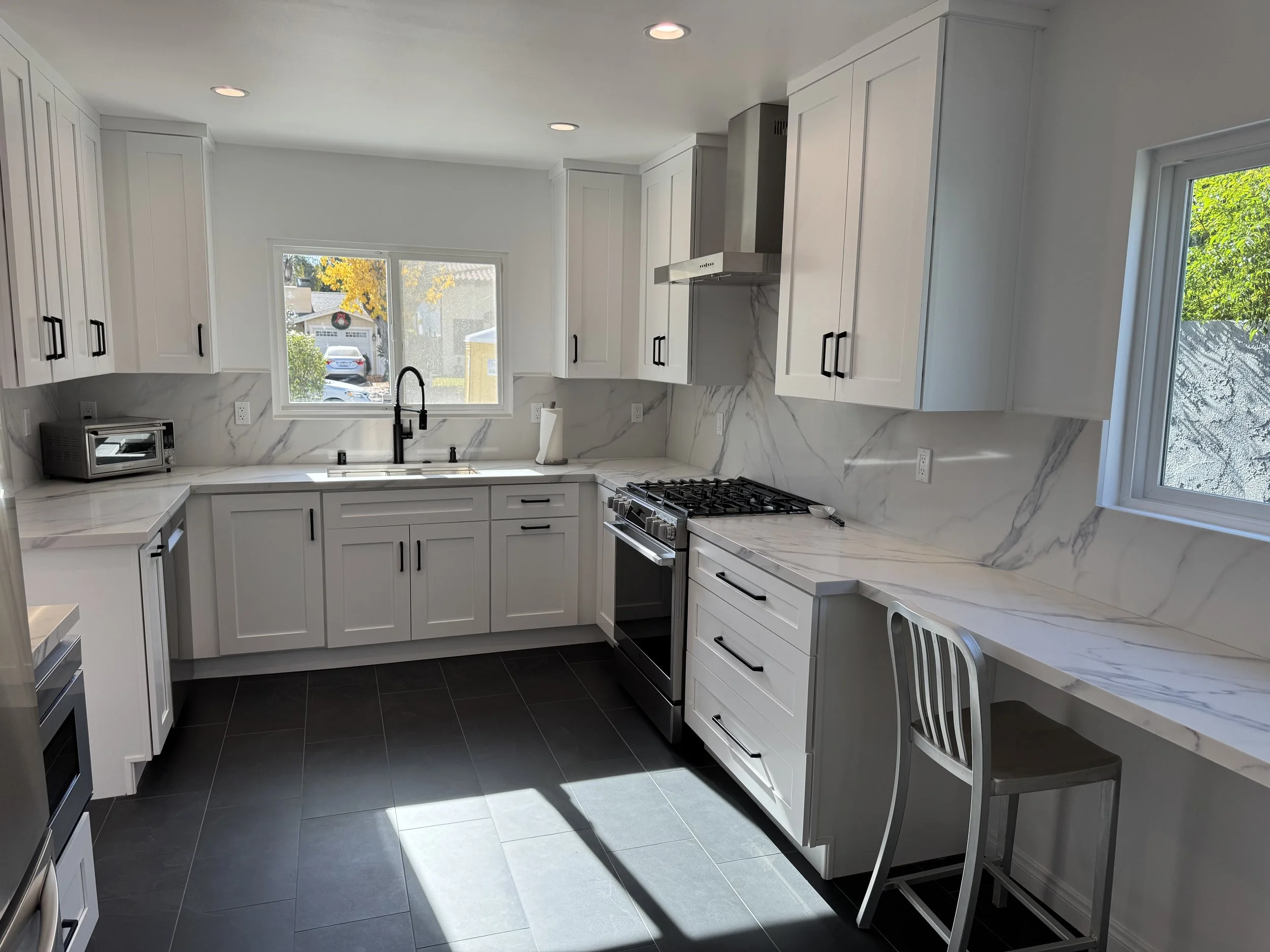 Modern kitchen with white cabinets, black handles, marble countertops, and a window above the sink showing an outdoor scene. There are mixed appliances, including a stove, microwave, and a small toaster oven. A chair is tucked under a marble countert