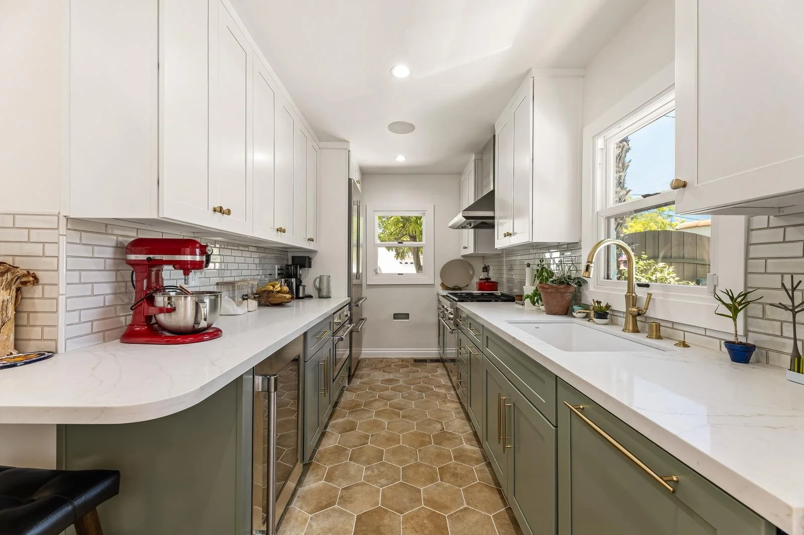 A modern kitchen with white upper cabinets, gray lower cabinets, and brass hardware. Contains a red stand mixer, potted plants, and a window above the sink with a view of trees and a fence.