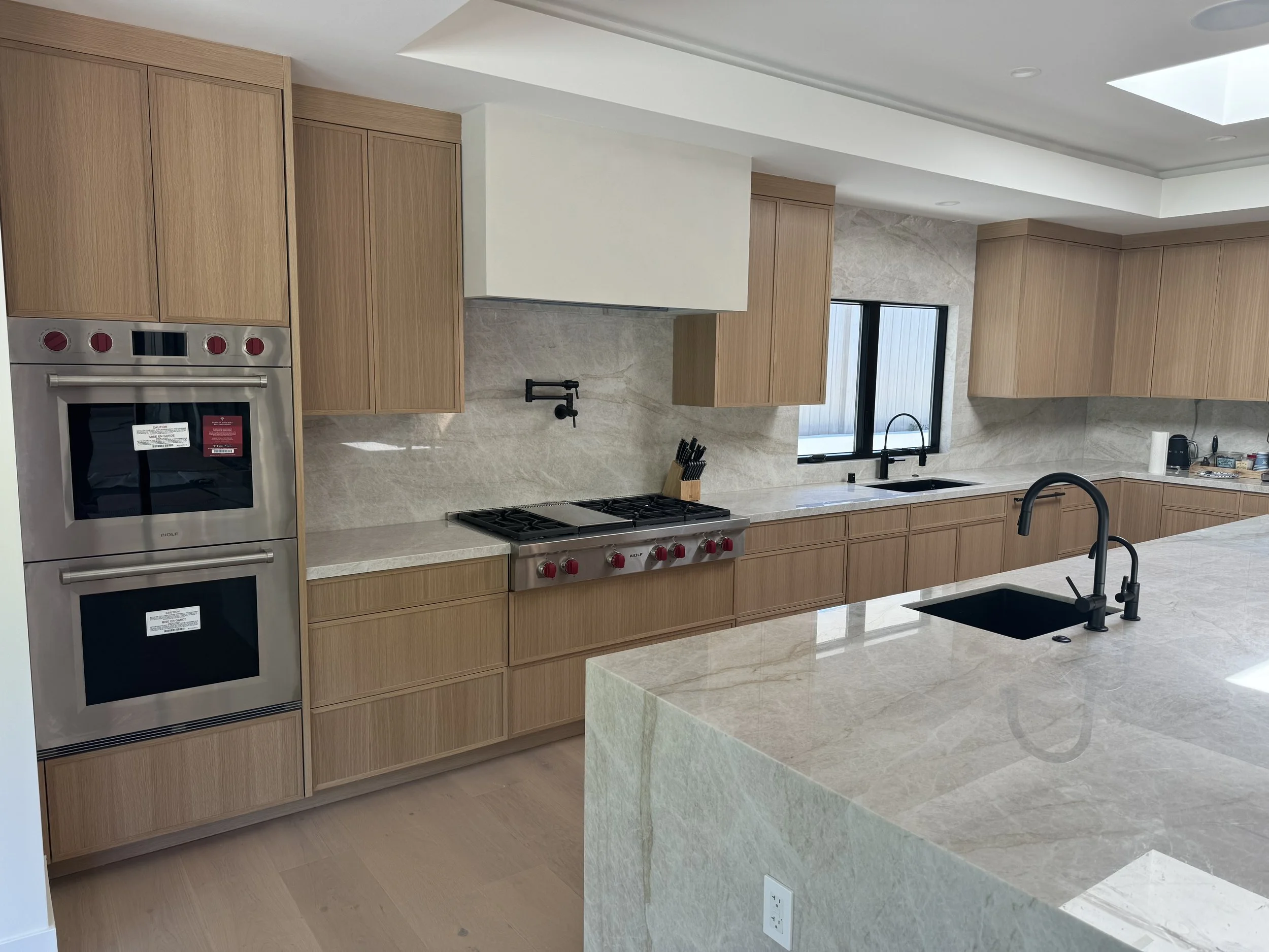 Modern kitchen with light wood cabinets, a kitchen island with a sink and black faucets, a double oven, a gas stove, a window above the sink, and various kitchen appliances and utensils.