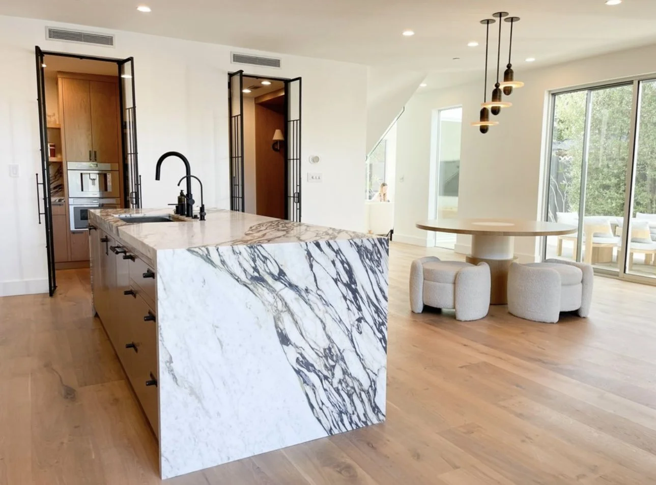 Modern open-concept kitchen and dining area with a marble kitchen island, light wood flooring, and sliding glass doors leading outside, featuring minimalistic lighting fixtures and cozy off-white seating.
