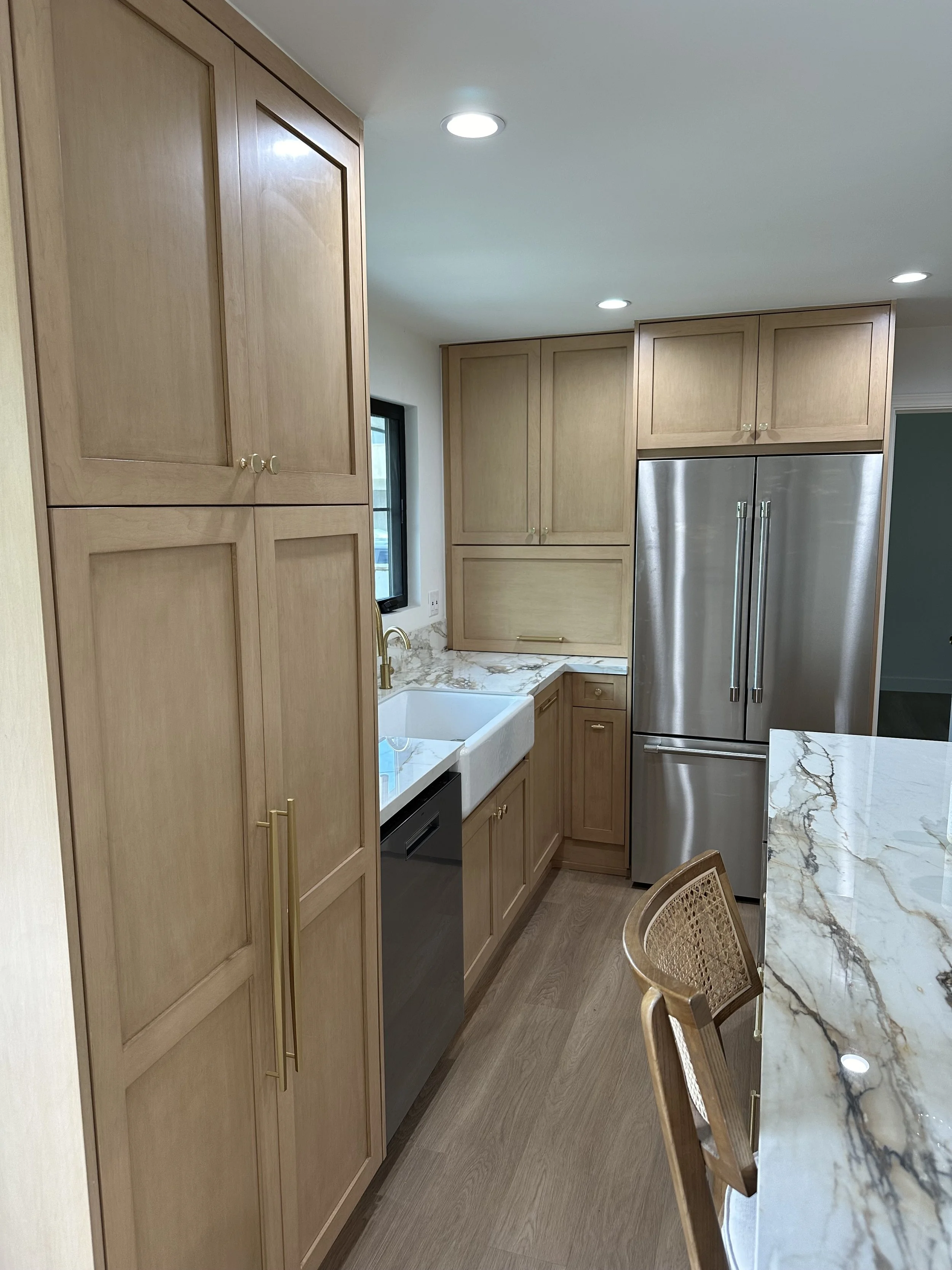 Modern kitchen with light wood cabinets, marble countertops, stainless steel refrigerator, front-facing sink, and wooden chair.