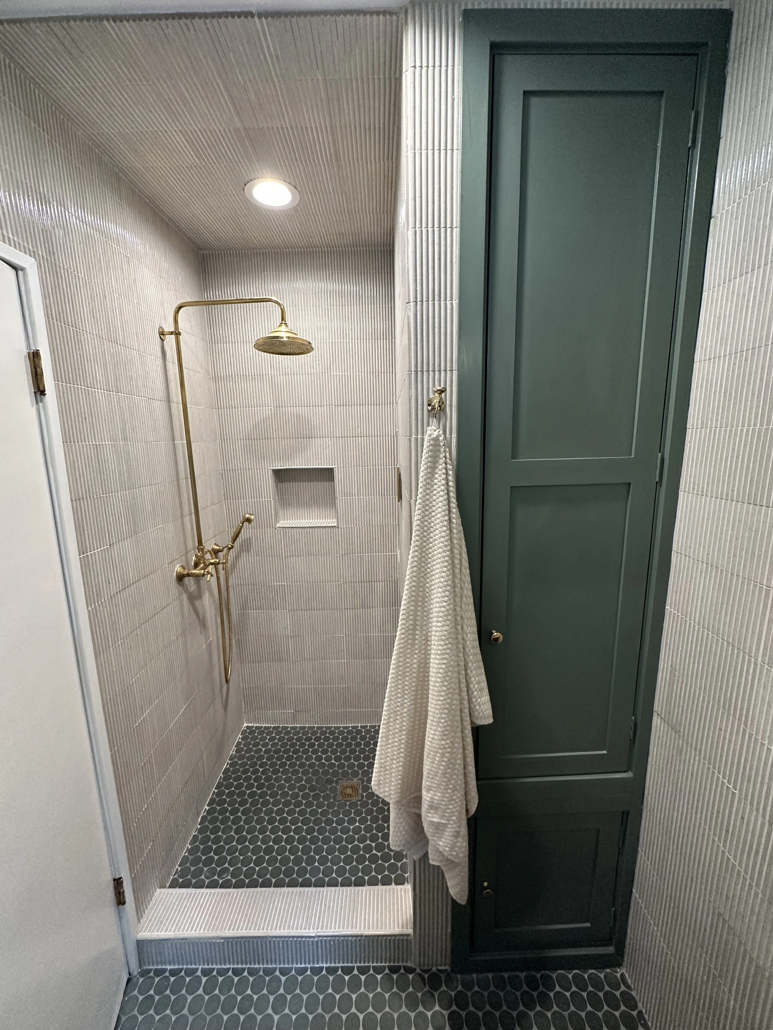 Small bathroom with a walk-in shower with brass fixtures, beige tile walls, black hexagonal floor tiles, a built-in niche, white towel hanging on a drilled hook, and dark green storage cabinets.