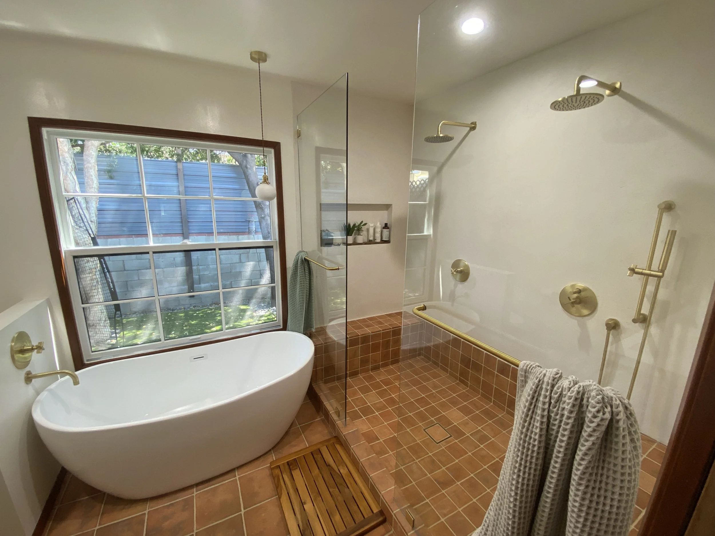 Bathroom with a large bathtub, a walk-in shower with multiple showerheads, and a large window with a view of trees and a blue fence.