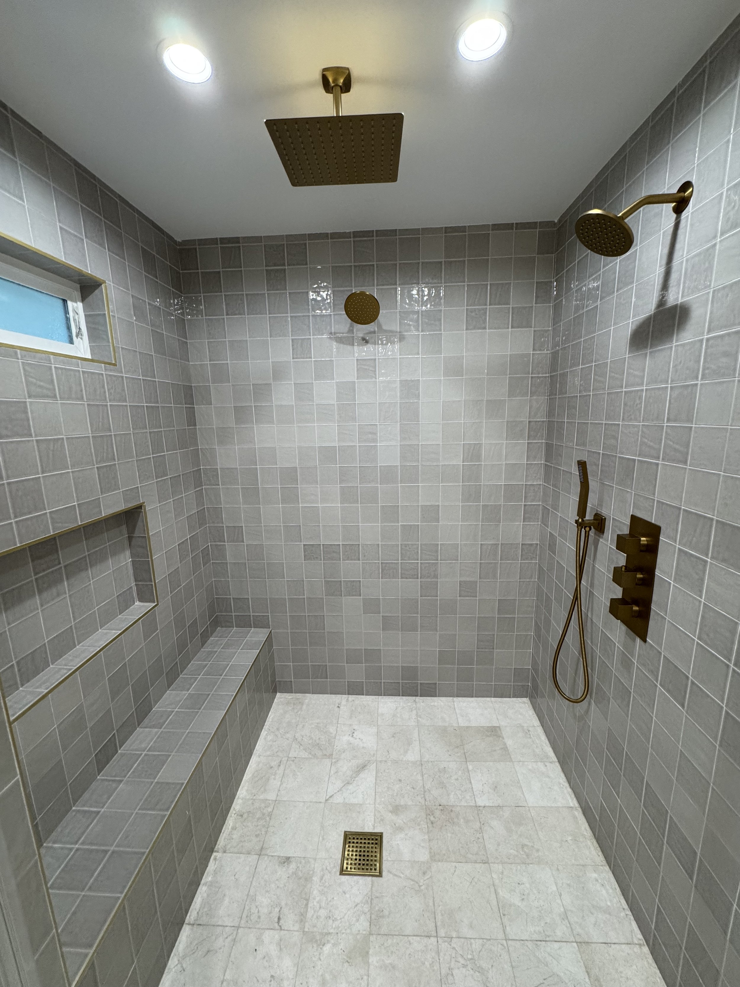 A modern walk-in shower with gray tiled walls, a built-in bench, three shower heads including a rain shower, a handheld shower, and two overhead spotlights, with a small window on the upper left.