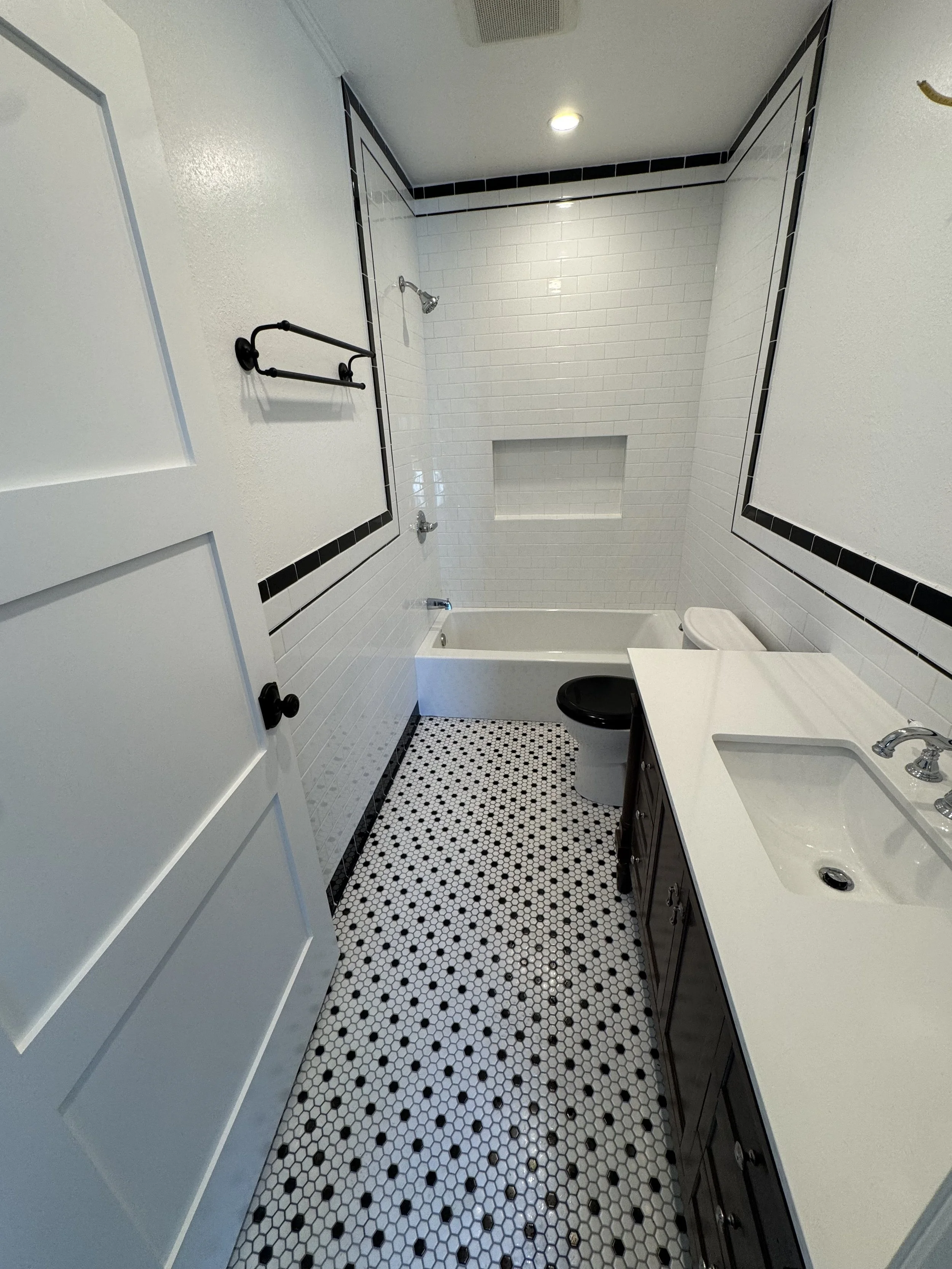 Bathroom with black and white tile floor, white tiled bathtub and shower, dark wood vanity, white countertop, white walls with black tile accents, and a recessed shower shelf.