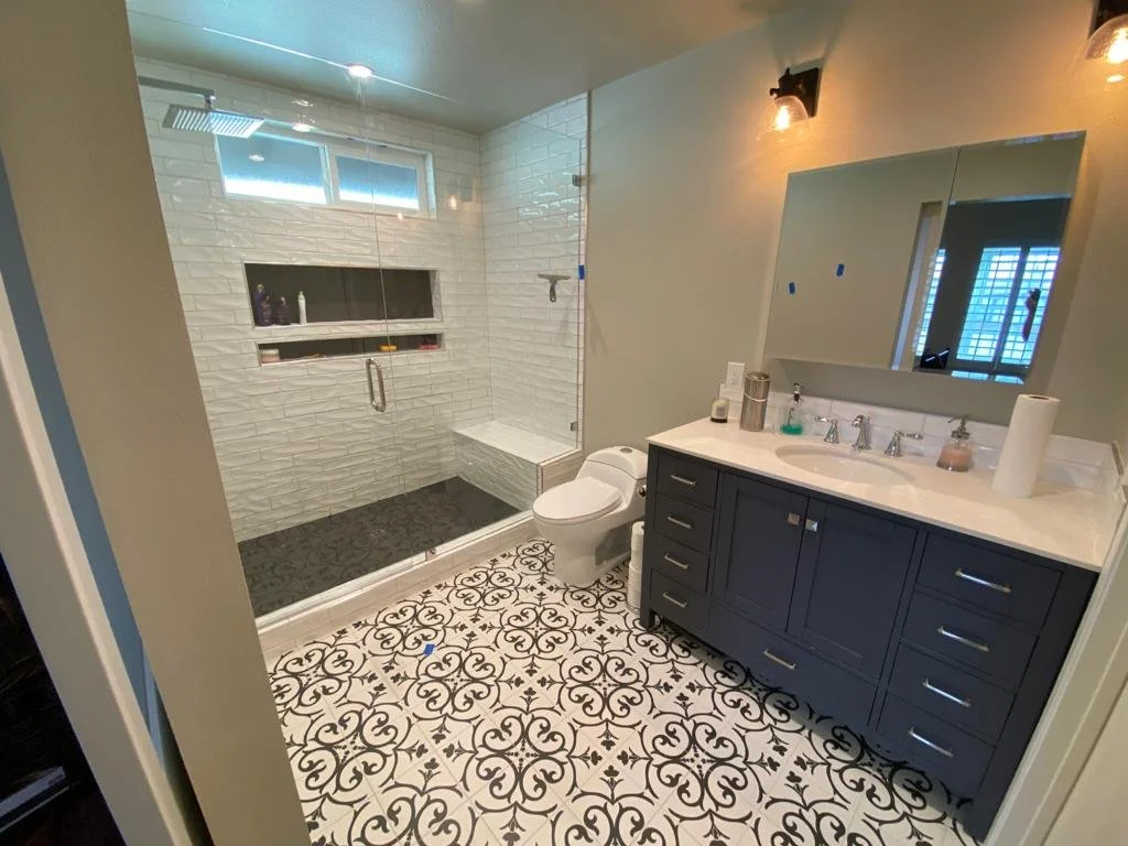 Modern bathroom with a walk-in glass shower, white subway tile walls, patterned black and white floor tiles, a dark blue vanity with drawers, a large mirror, and a toilet.