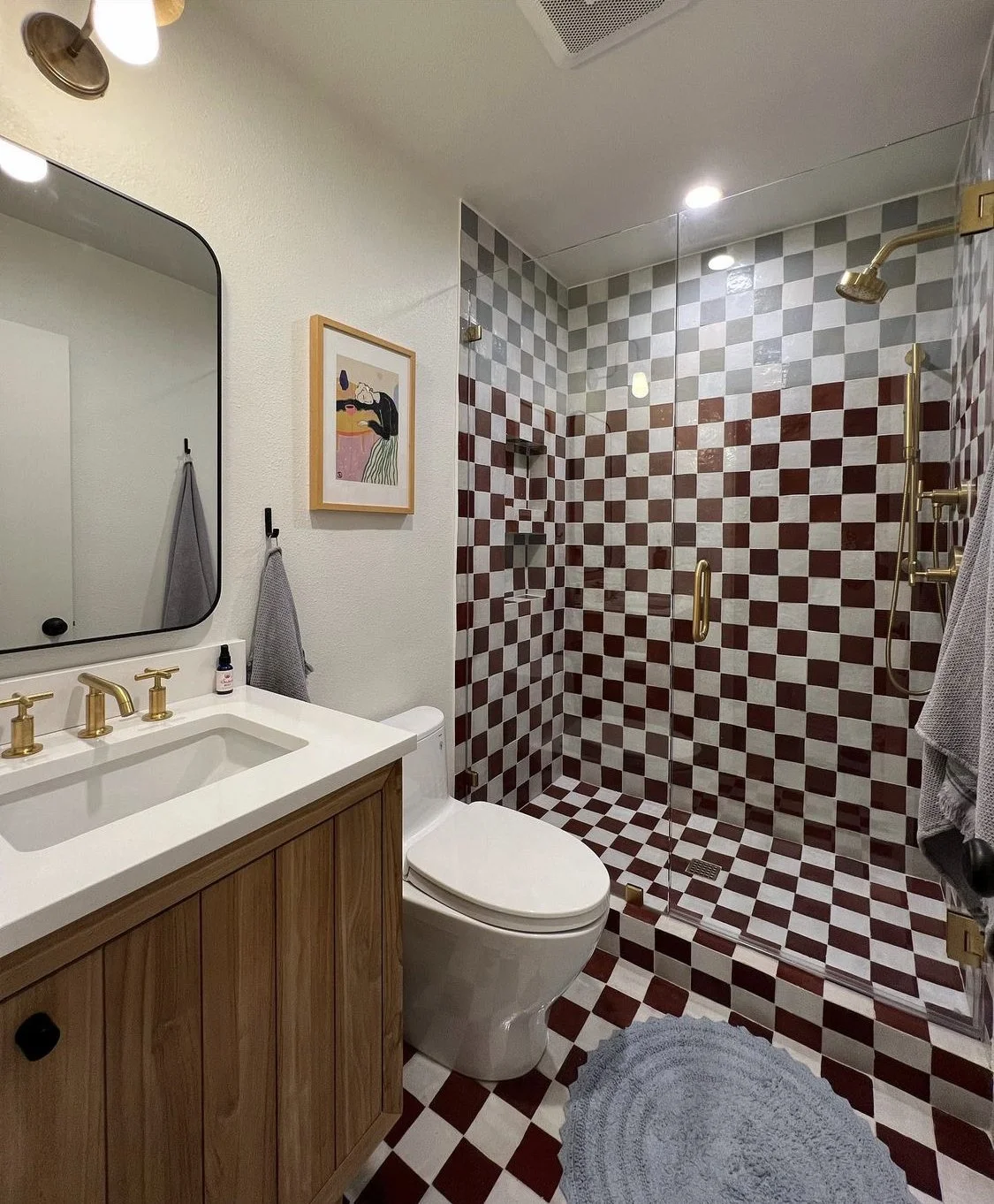 A modern bathroom with a checkered tile pattern in red and white in the walk-in shower, a white toilet next to a wooden vanity with gold fixtures, a mirror above the sink, and a small piece of colorful artwork on the wall.