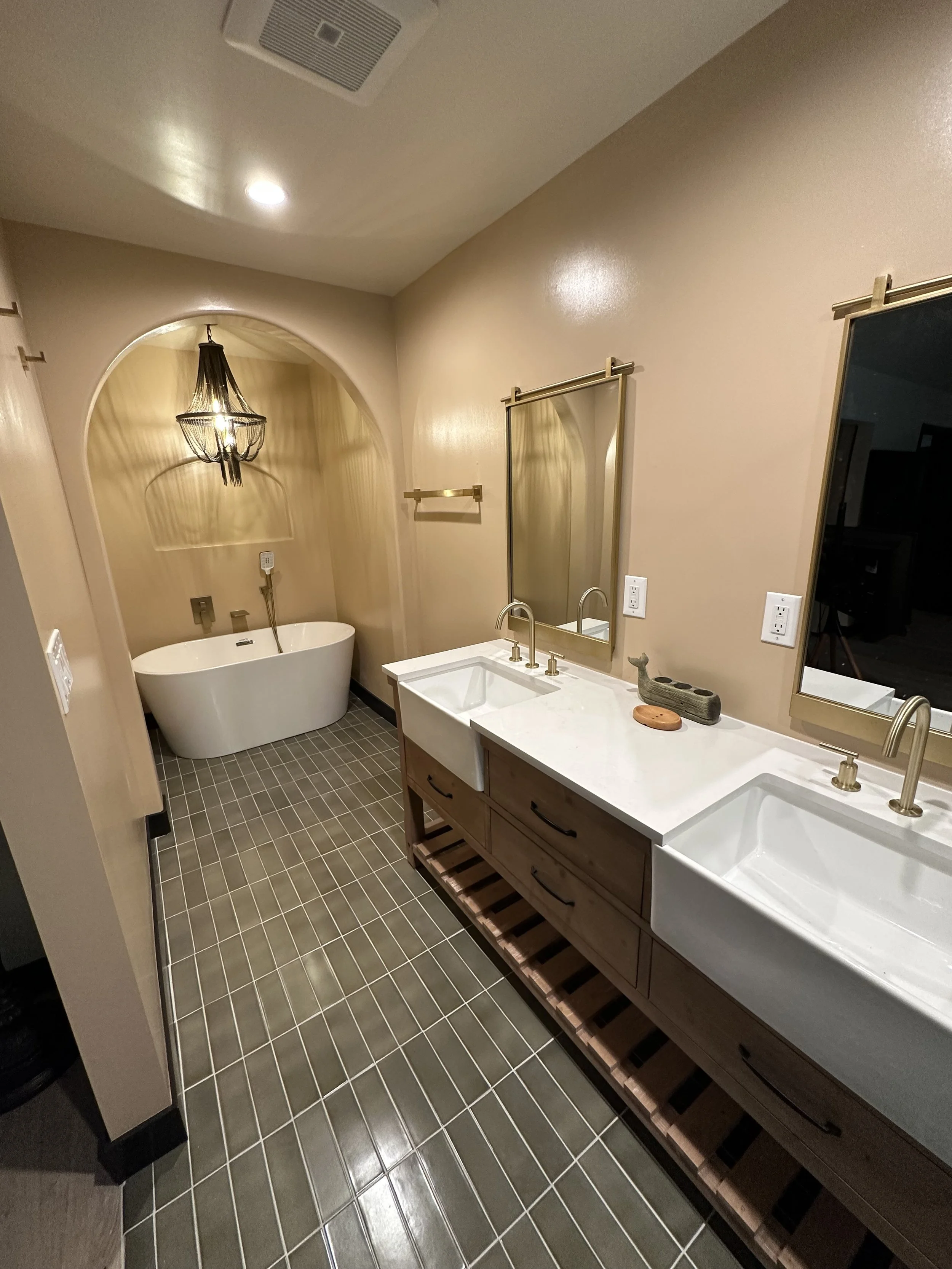 Bathroom with dual sinks, large mirror, wooden vanity, gray tile floor, and a bathtub with a chandelier overhead.
