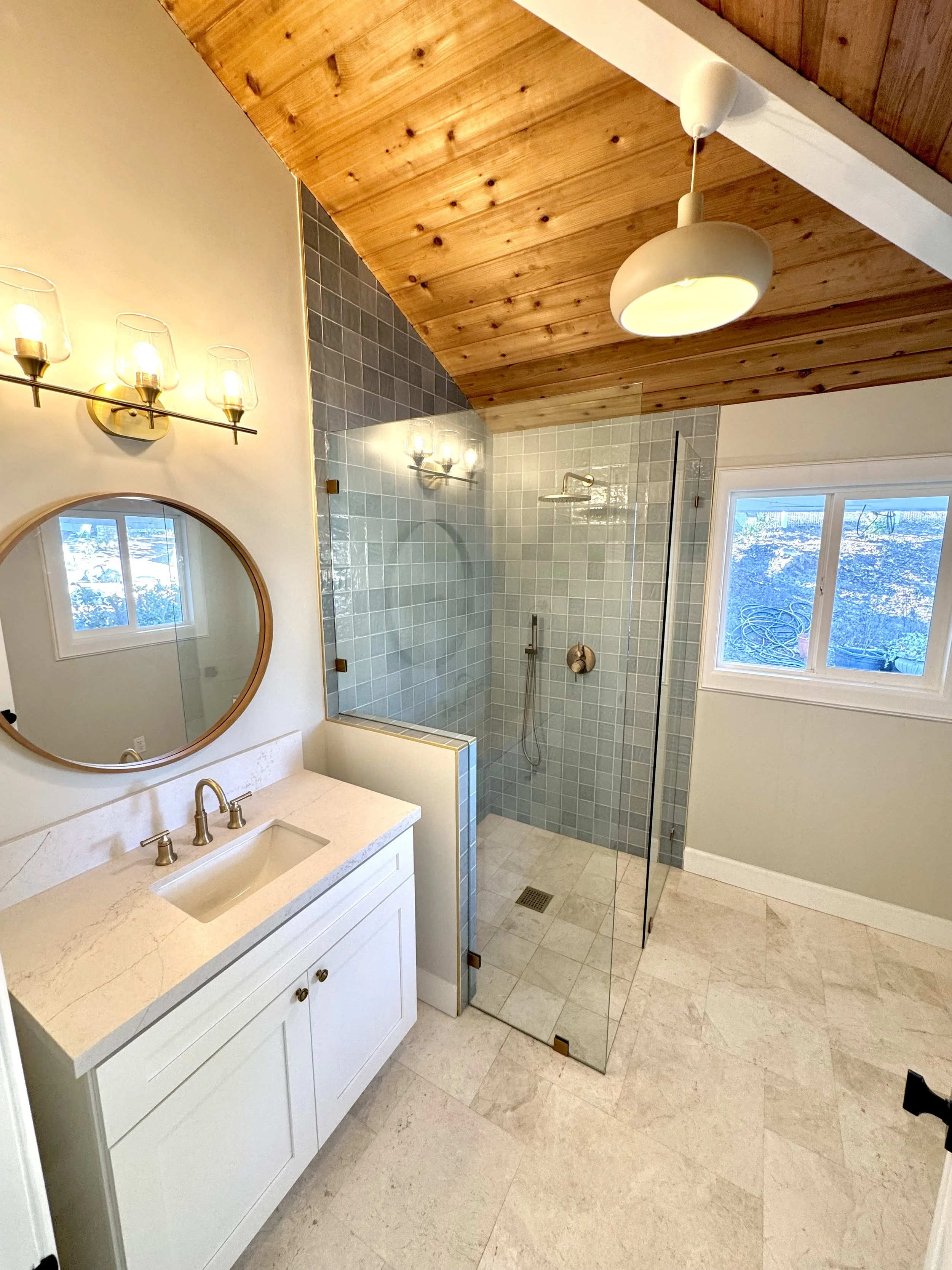 Modern bathroom with white vanity, round mirror, light fixture with four glass shades, glass shower enclosure with light blue tiles, wood-paneled ceiling, window showing outdoor landscape.