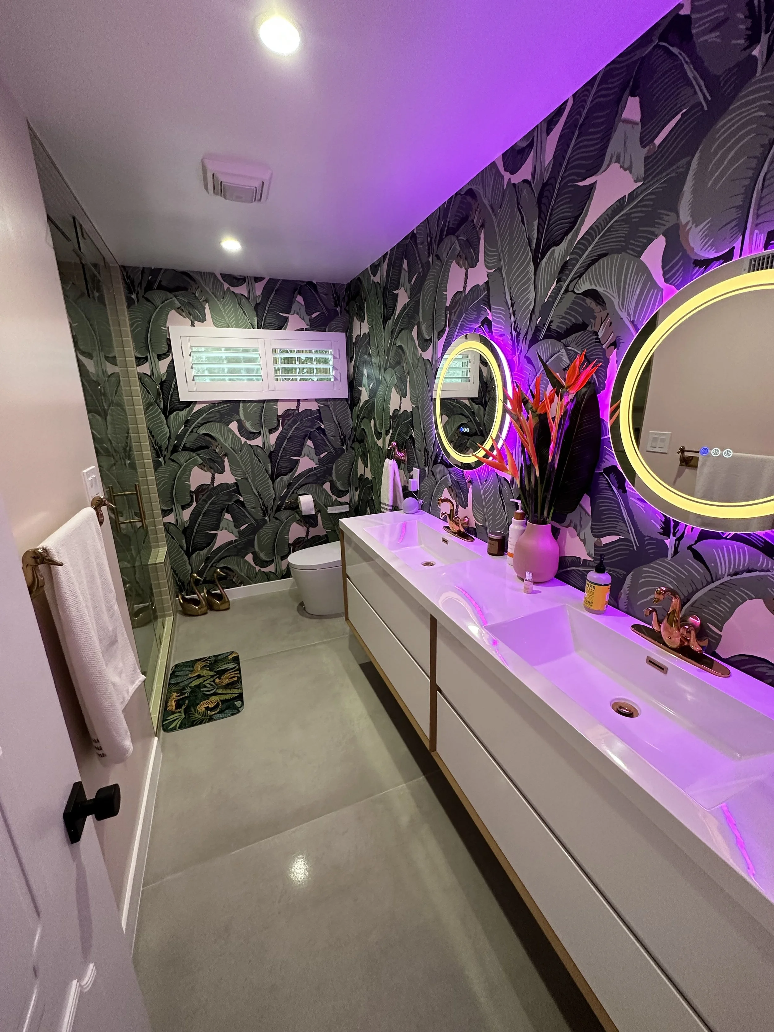 Modern bathroom with tropical leaf wallpaper, illuminated round mirrors with LED lights, double sink vanity with decorative golden unicorn figurines, large plant in a white vase, and purple LED lighting.
