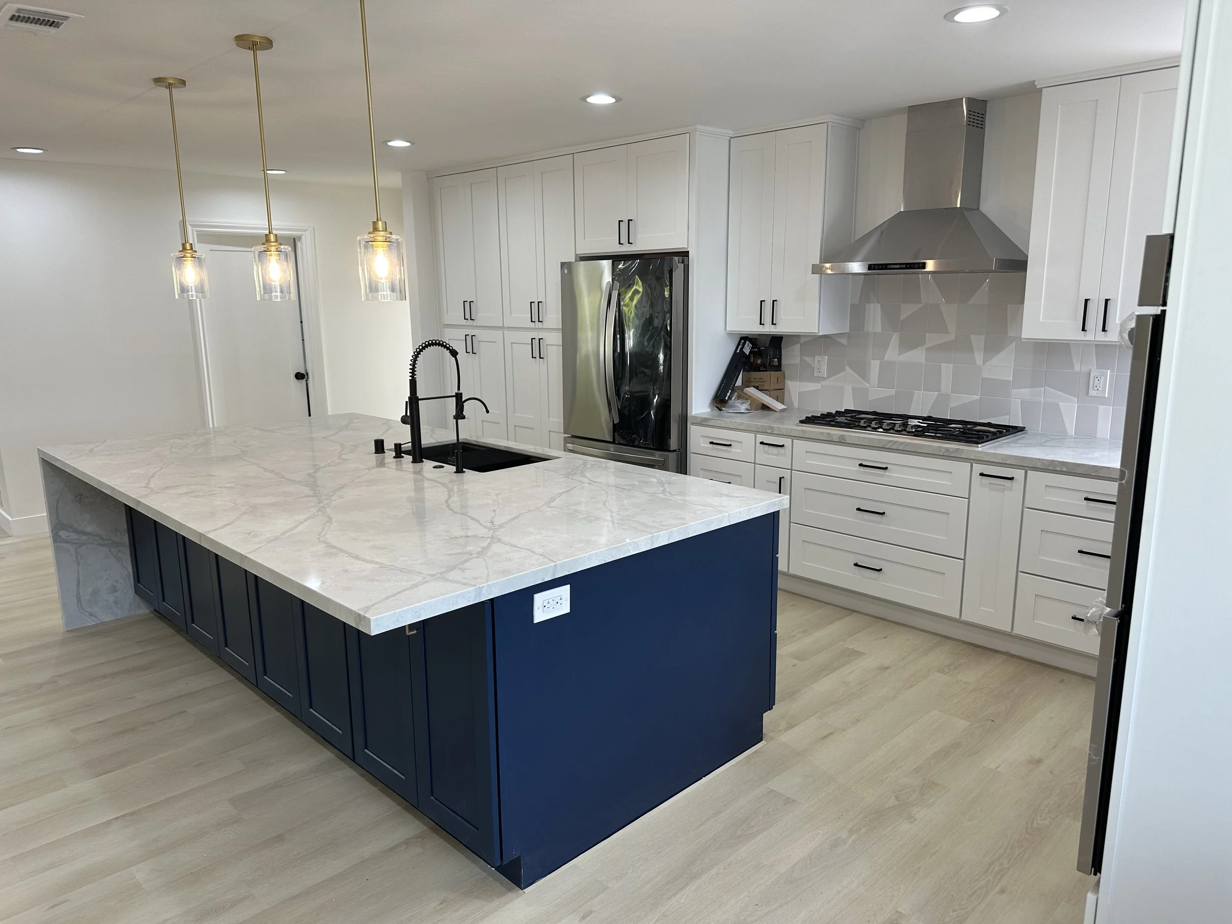 Modern kitchen with white cabinets, a large island with a blue base, marble countertop, black sink, black faucet, and three pendant lights, stainless steel refrigerator, range hood, and geometric tile backsplash.