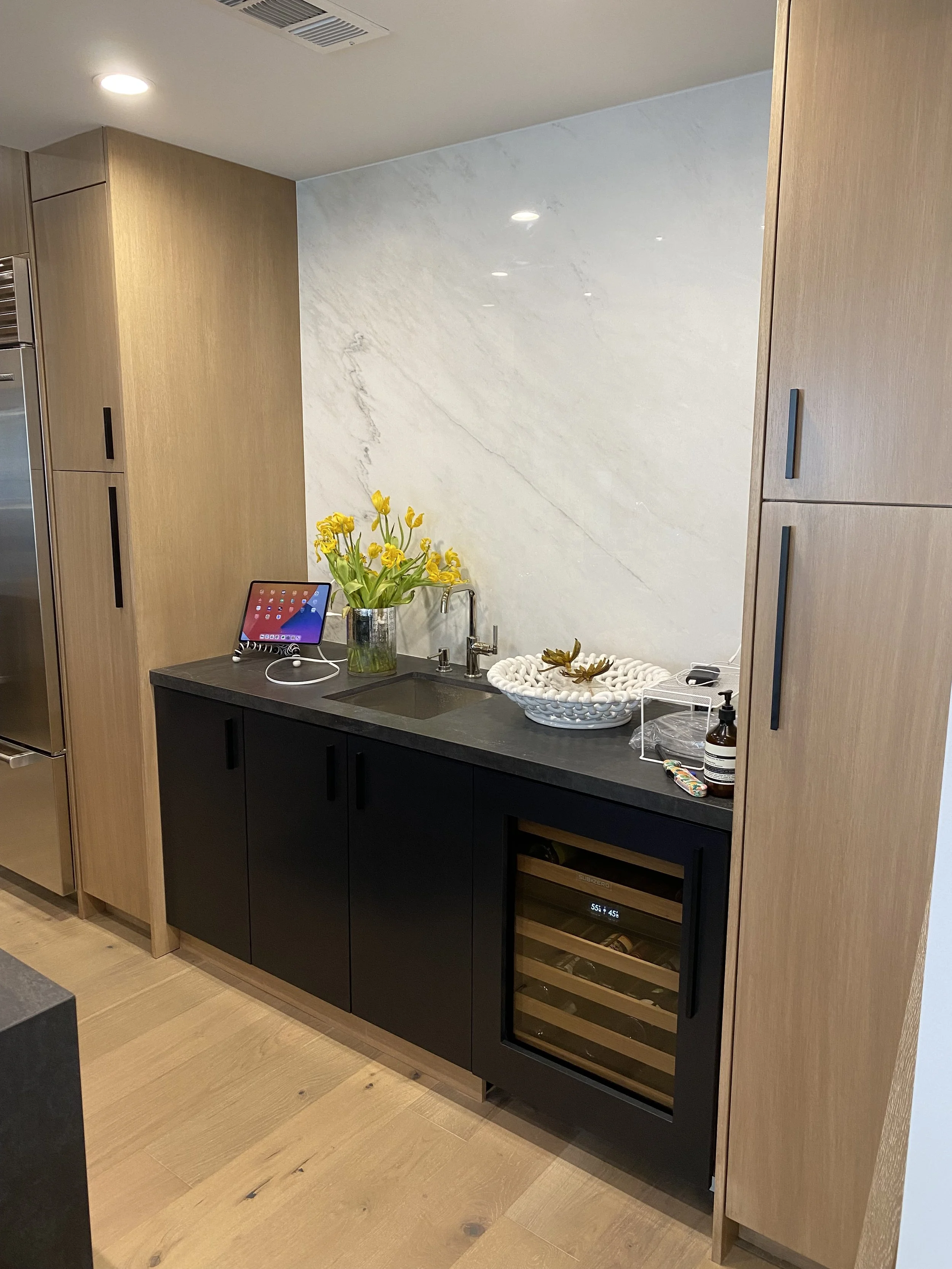 Kitchen with black cabinets, wooden built-in cabinets, marble backsplash, small sink, vase of yellow flowers, white decorative bowl, and small appliances.