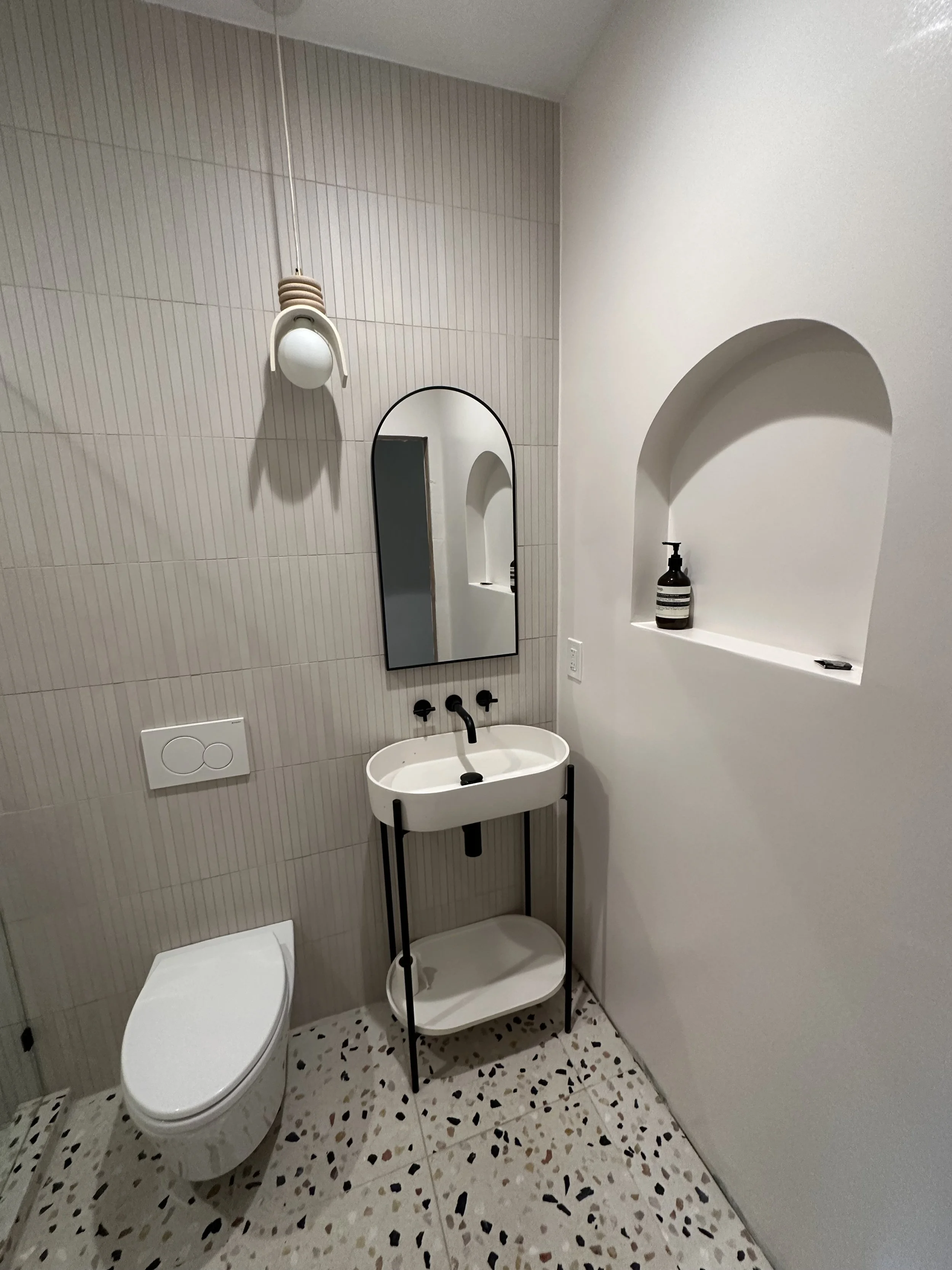 A small modern bathroom with a wall-mounted toilet, a small white sink with a black stand, a vertical oval mirror, peach-colored vertical tiles on the wall, a wall niche with a brown bottle, and a terrazzo floor.