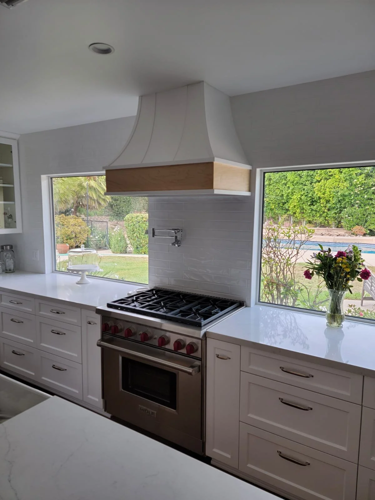 Modern kitchen with white cabinets, a built-in stainless steel oven, a gas stovetop, a white range hood, windows showing outdoor greenery, and a vase of flowers on the countertop.