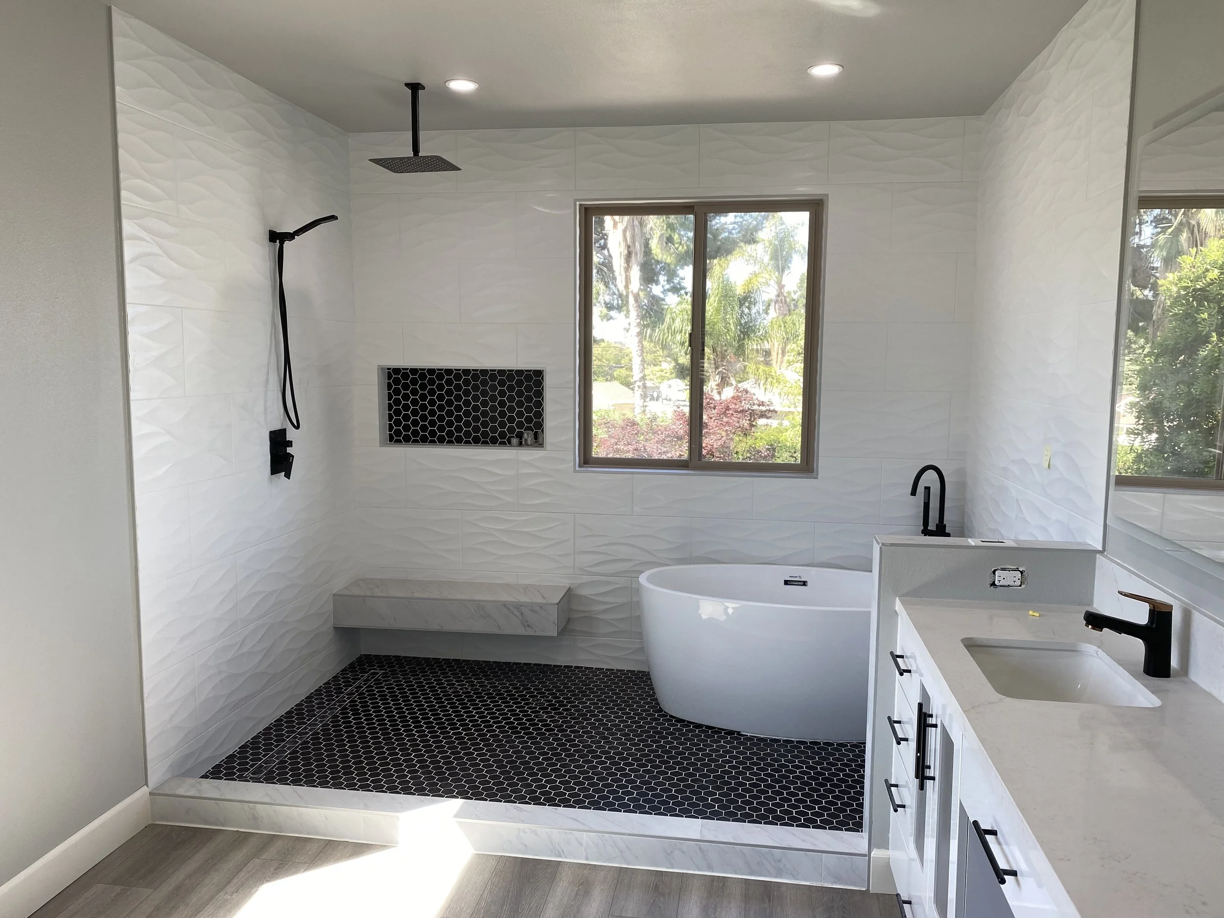 Modern bathroom with walk-in shower featuring black fixtures, a freestanding bathtub, a window with trees outside, and a white vanity with black handles.
