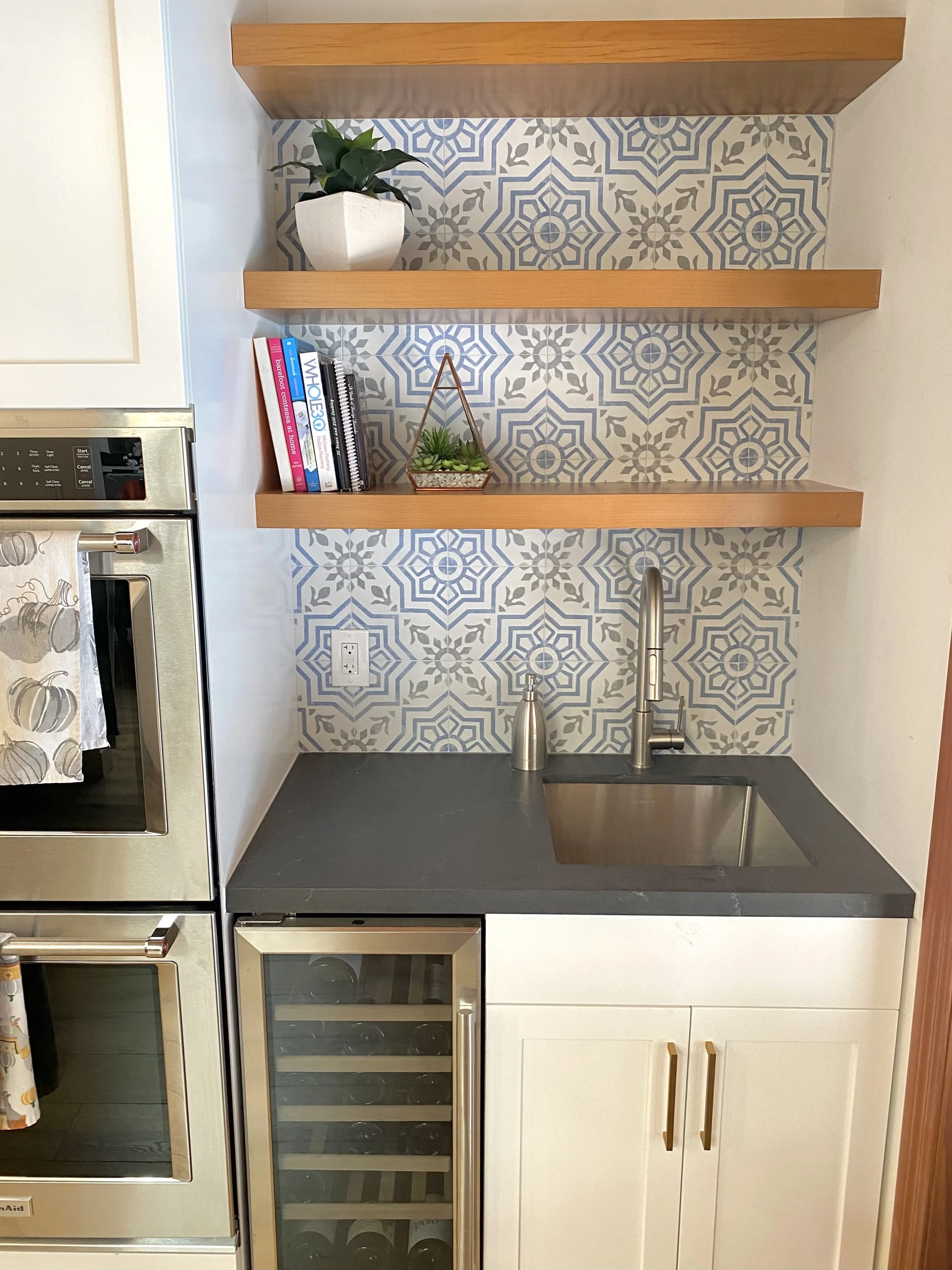 Kitchen corner with two wooden shelves holding a white potted plant, books, and a small succulent; gray countertop with a built-in sink and modern faucet; patterned backsplash and a wine cooler below the counter.