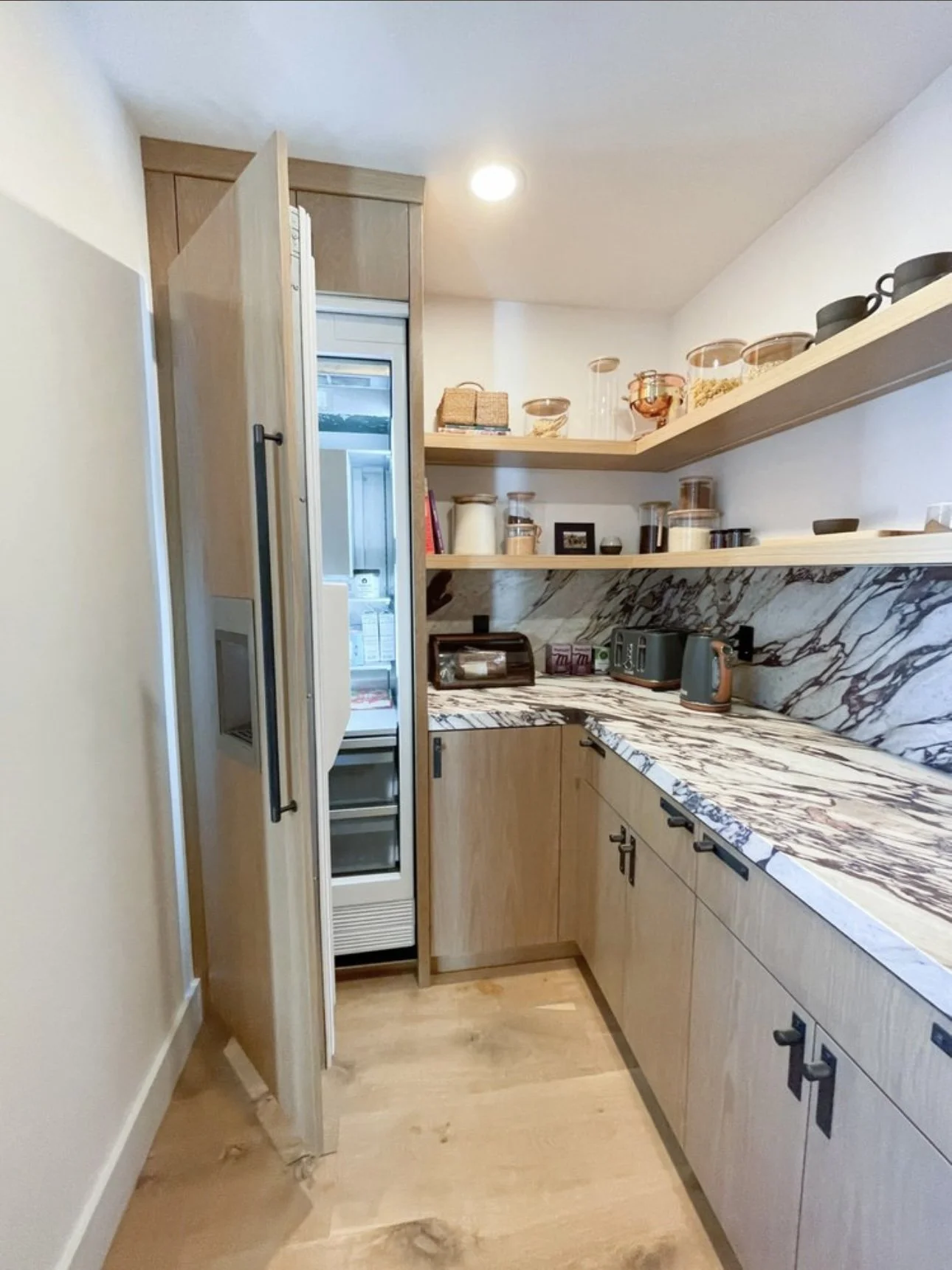 A small modern kitchen with a marble countertop and open wooden shelves holding glass jars and dishes. A built-in refrigerator with a door open reveals shelves inside. The cabinets are light wood, and the floor is light hardwood. There's a toaster, k