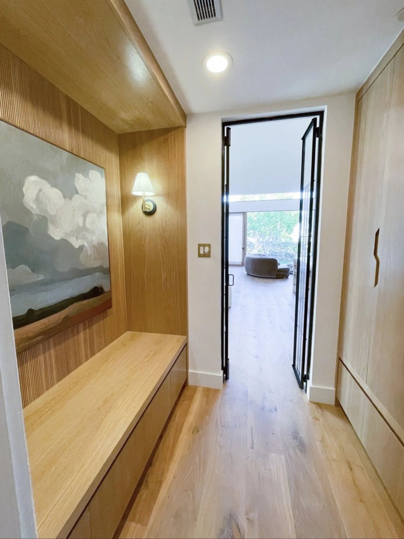 A hallway with wooden flooring, a wooden bench on the left, a wall-mounted lamp, a painting, and a door leading to a room with a sofa and large window showing trees outside.