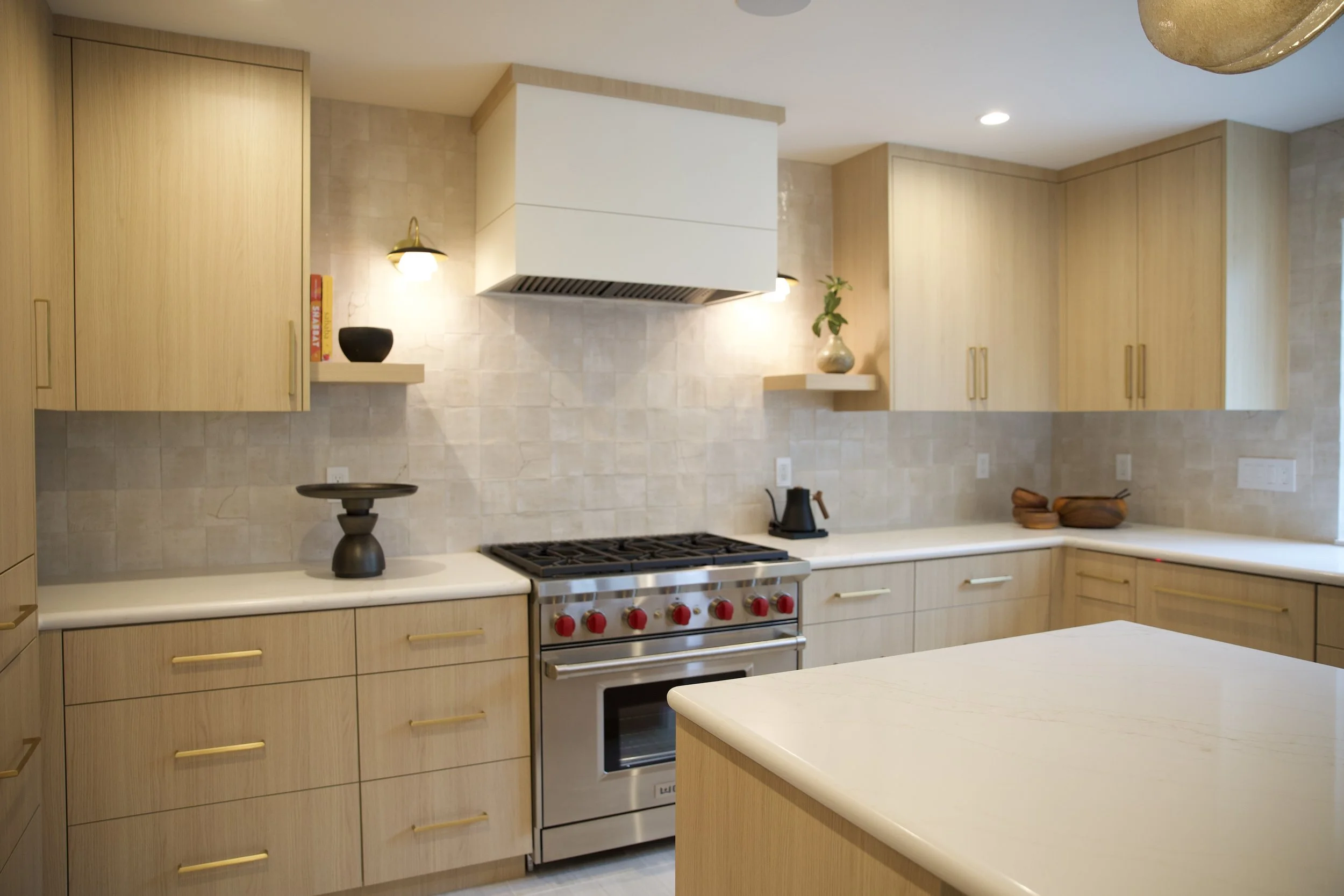 Modern kitchen with light wood cabinets, stainless steel stove with red knobs, white countertops, beige tiled backsplash, and decorative items on shelves.