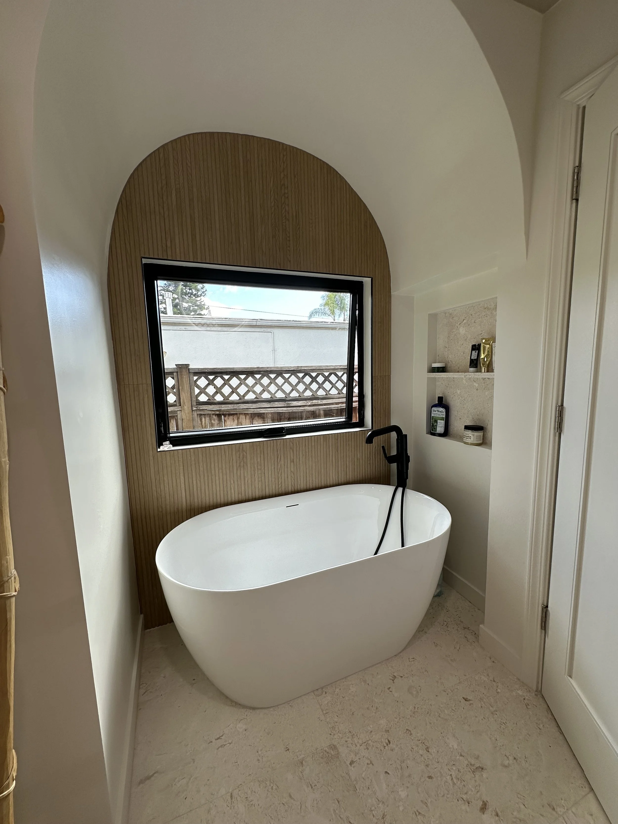 Modern bathroom with a freestanding bathtub below a large window, featuring a wooden accent wall and built-in shelves to the right.