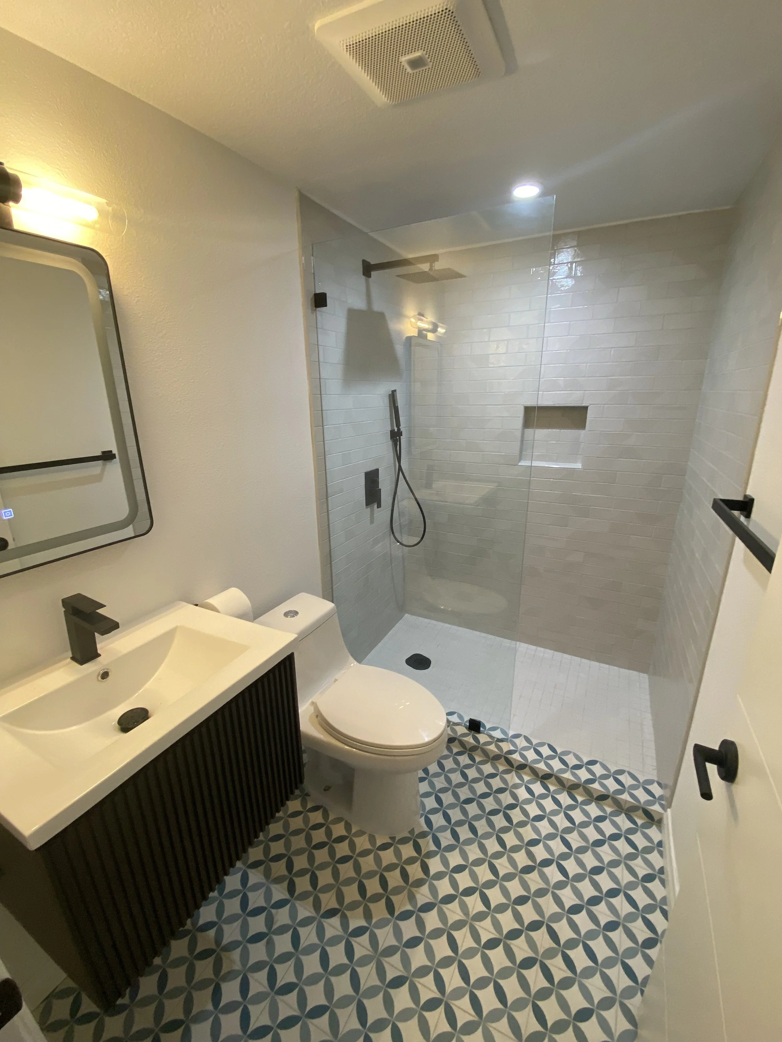 Modern bathroom with patterned tile floor, white toilet, black vanity with sink, wall mirror, and a walk-in shower with glass partition and white subway tiles.