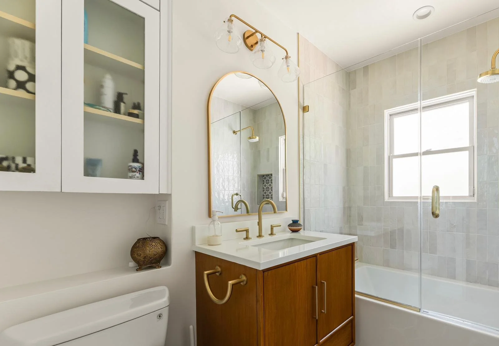 Modern bathroom with wooden vanity, gold fixtures, large mirror, glass shower enclosure, window, and white toilet.