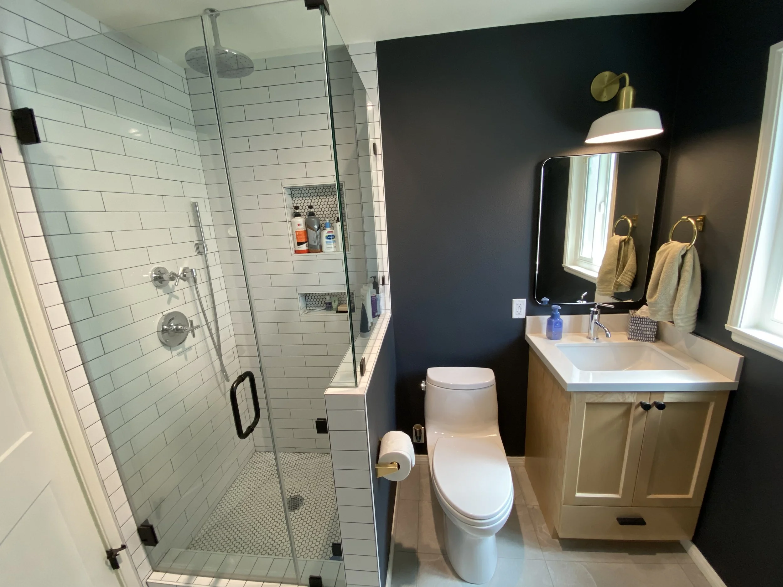Modern bathroom with white tiled shower, black wall, wooden vanity with white sink, mirror, and chrome fixtures. Toilet with toilet paper holder, towel racks with towels, and two windows letting in natural light.