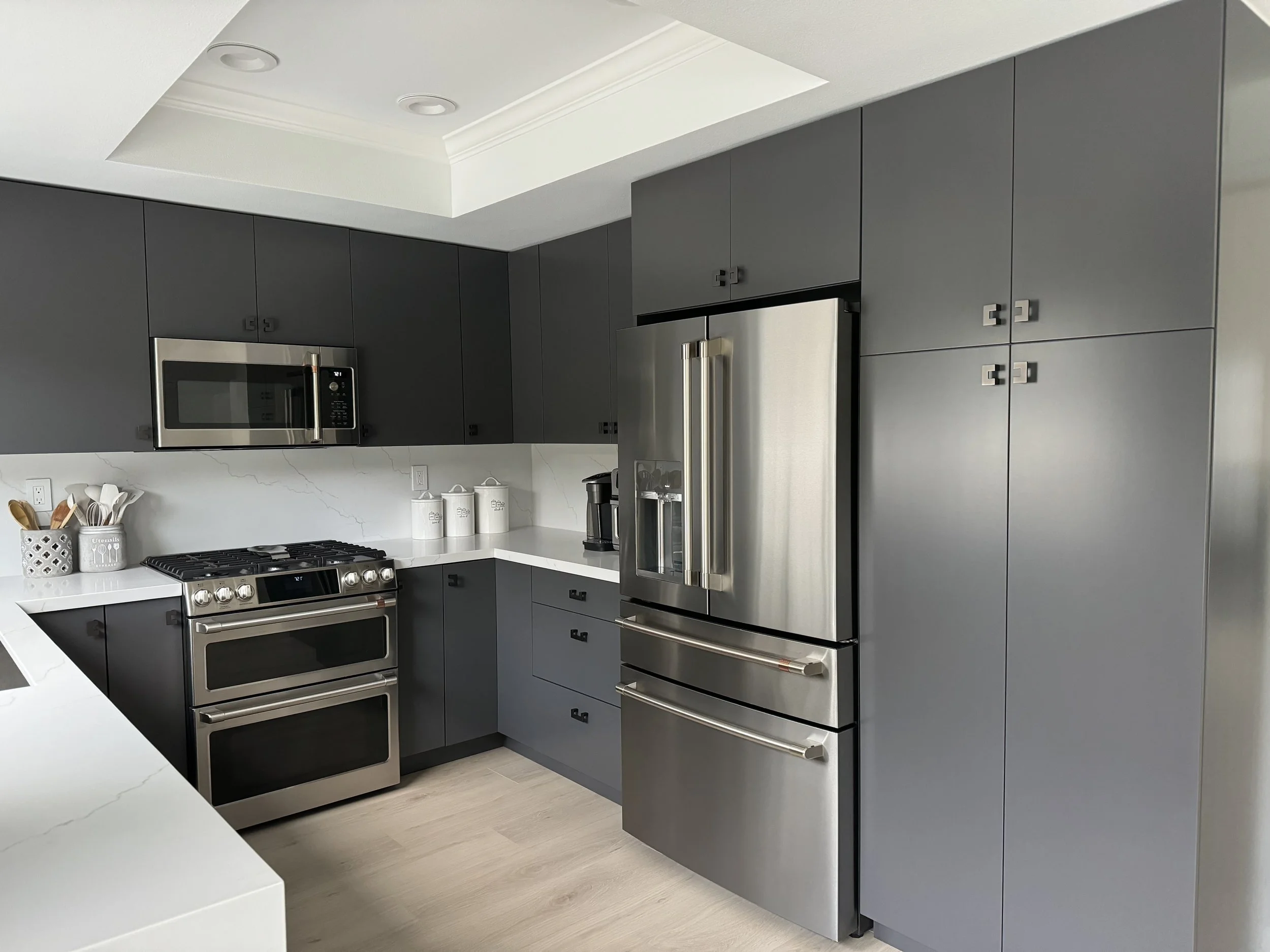Modern kitchen with gray cabinets, stainless steel appliances, including a double oven, microwave, and refrigerator, white marble countertops, and light wood flooring.