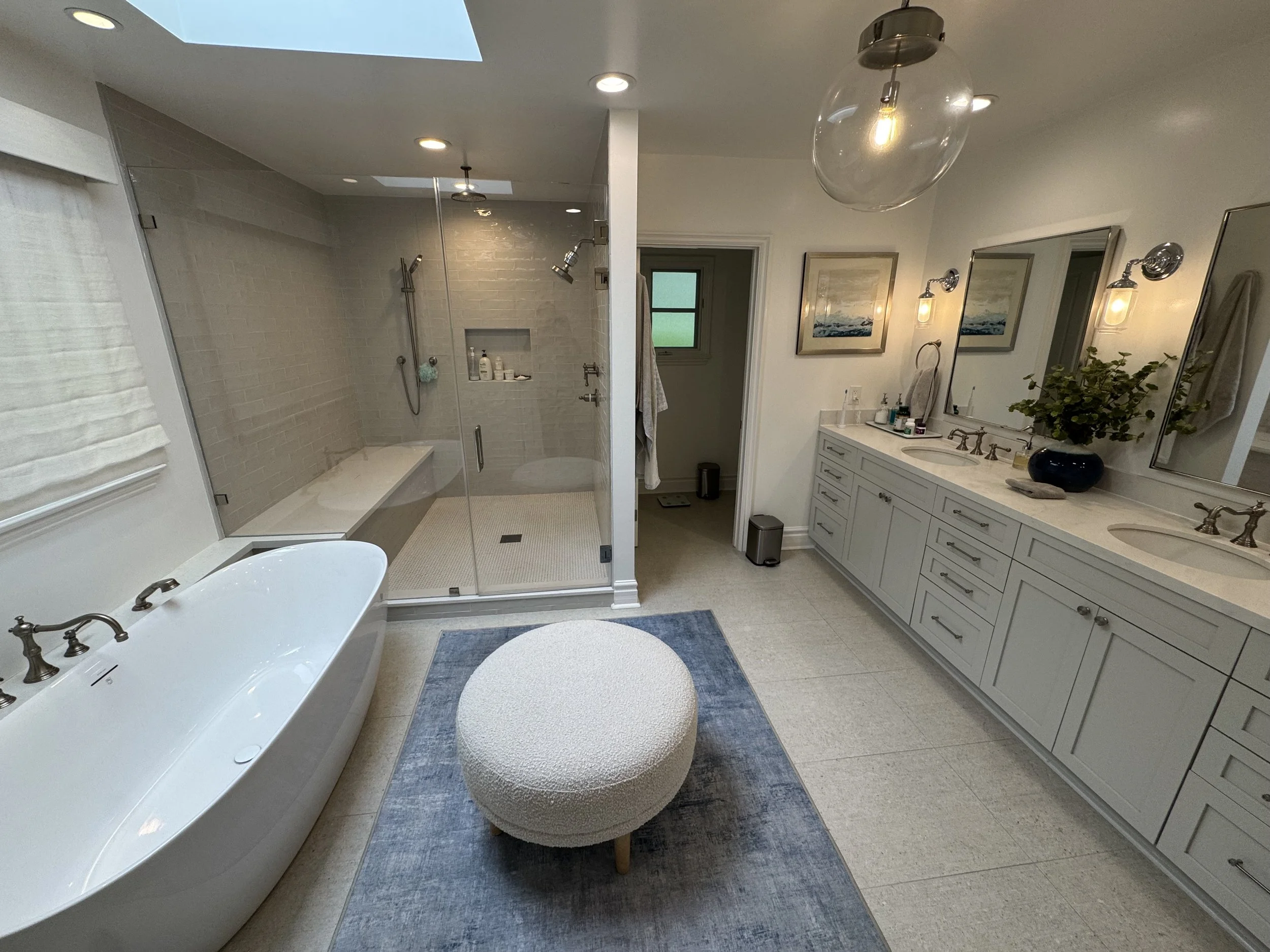 A spacious bathroom with a freestanding bathtub, a walk-in shower with a glass door, a double vanity with two mirrors, and modern lighting fixtures. The room features light-colored cabinets and a blue rug, with artwork and a plant on the vanity. A wi