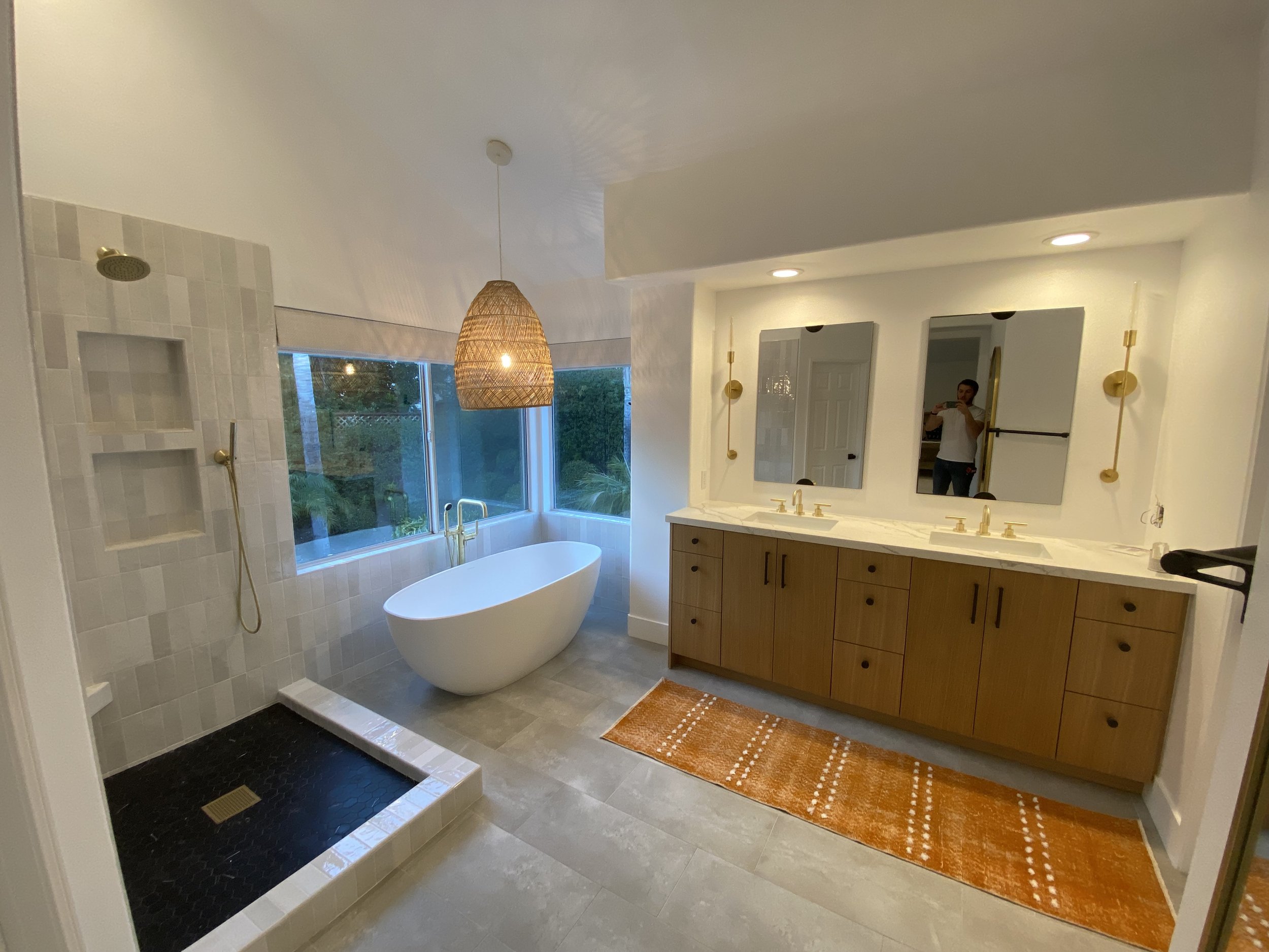 Modern bathroom with standalone bathtub near window, walk-in shower, double vanity with mirrors, and orange rug.