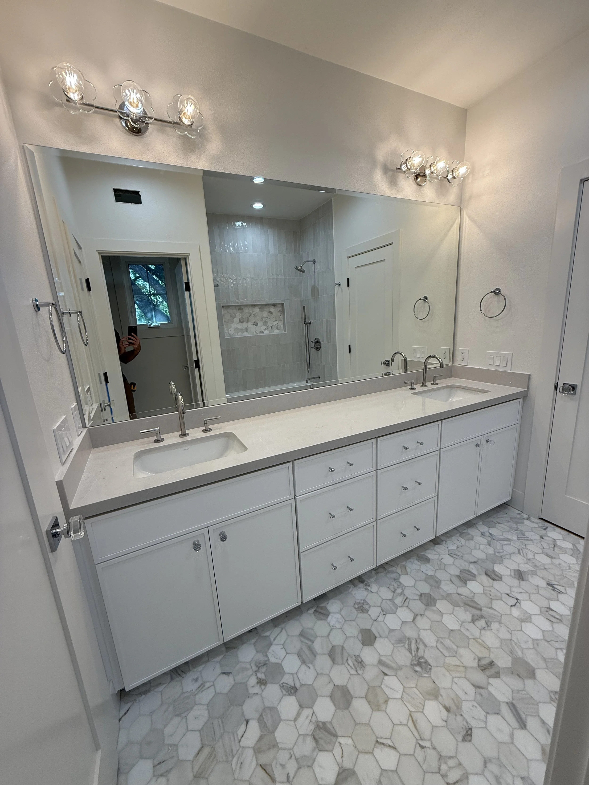 Modern bathroom with a large mirror above a white double vanity with a marble countertop and two sinks, illuminated by two wall-mounted light fixtures. The bathroom features a walk-in shower visible in the mirror’s reflection with glass door, showerh