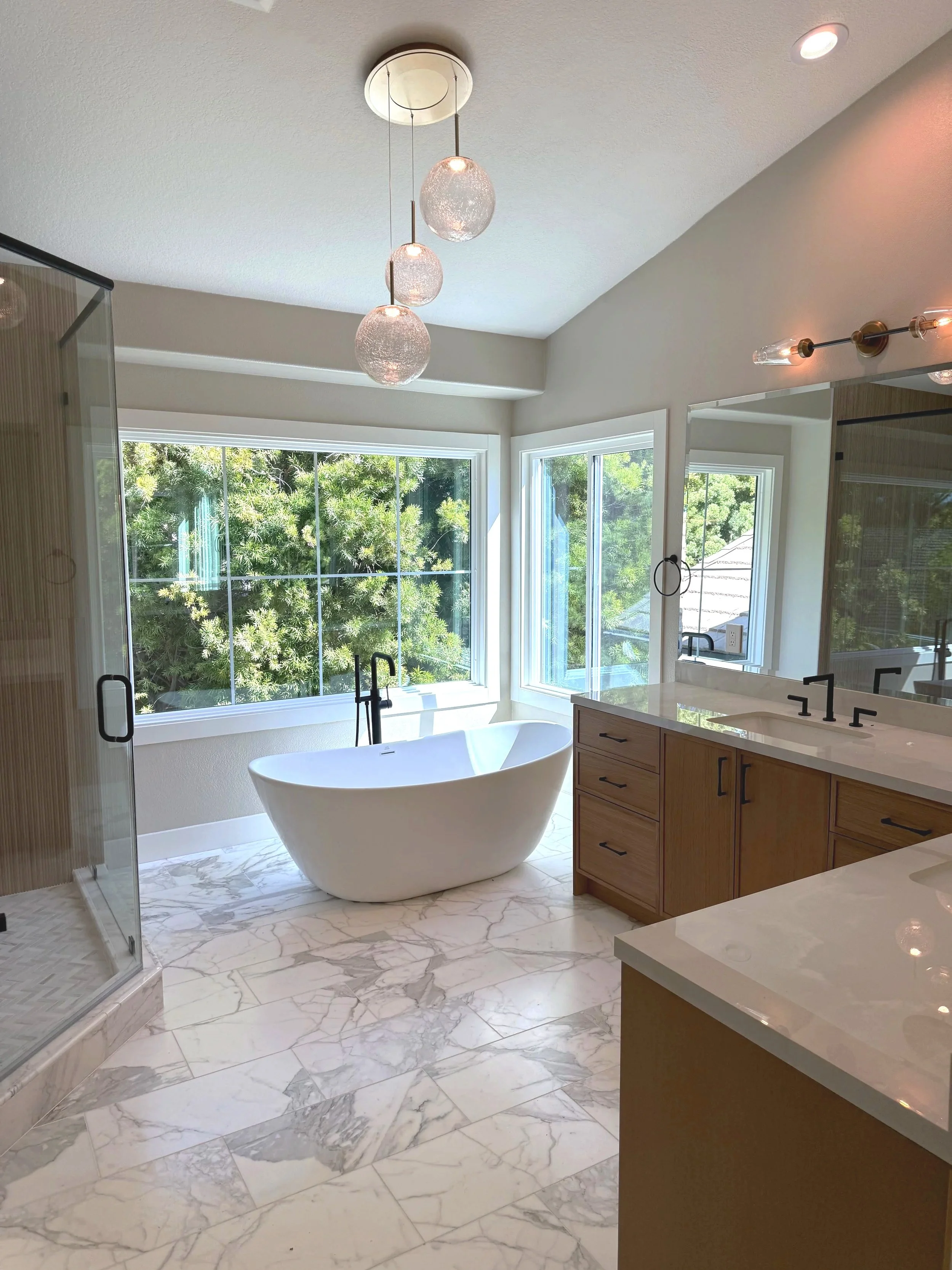 Modern bathroom with a white bathtub, marble floor tiles, wooden vanity with black handles, large window with trees outside, and a glass shower stall.