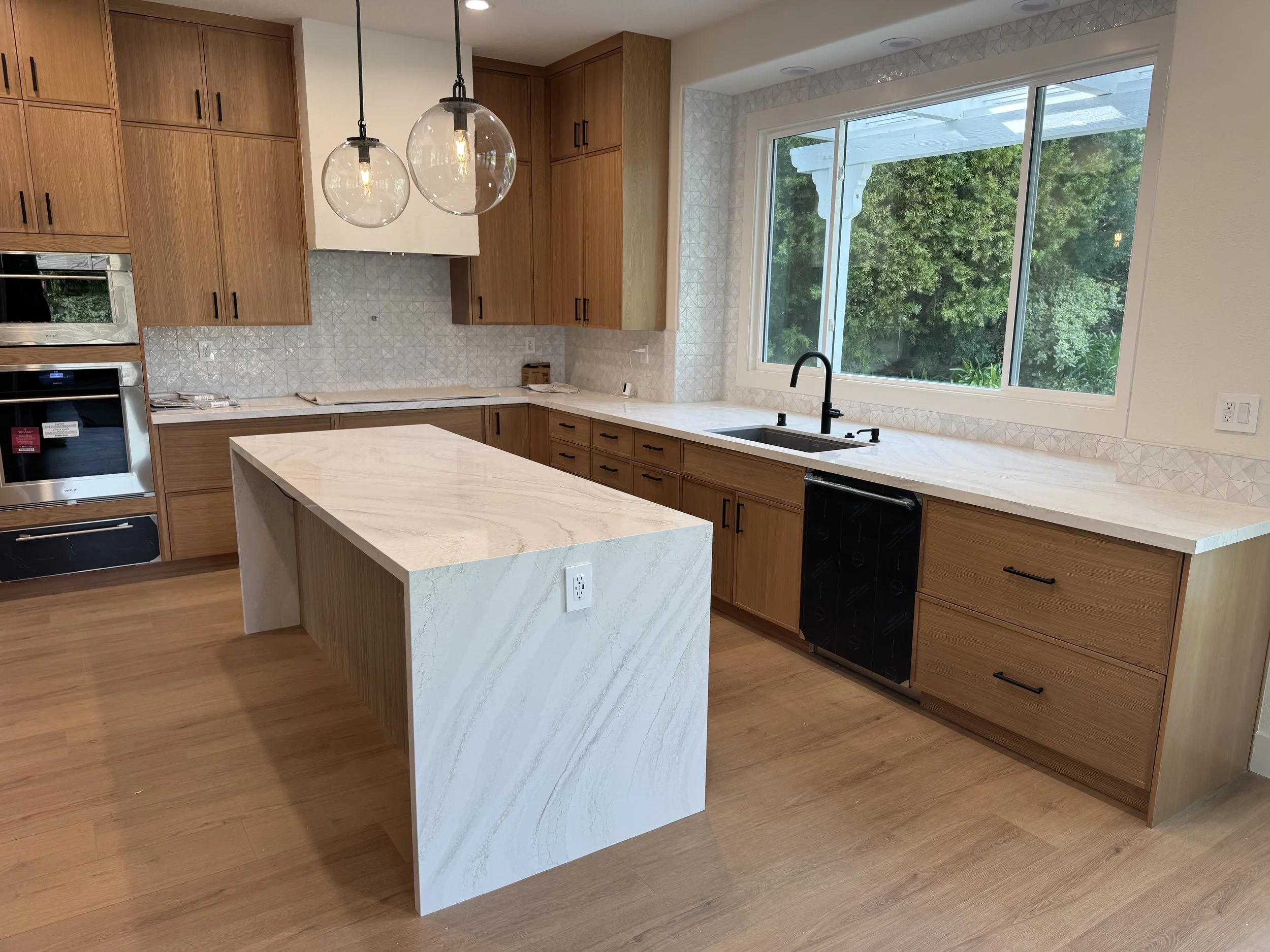 Modern kitchen with wooden cabinets, white countertop island, large window over sink, black faucet, and pendant lights.