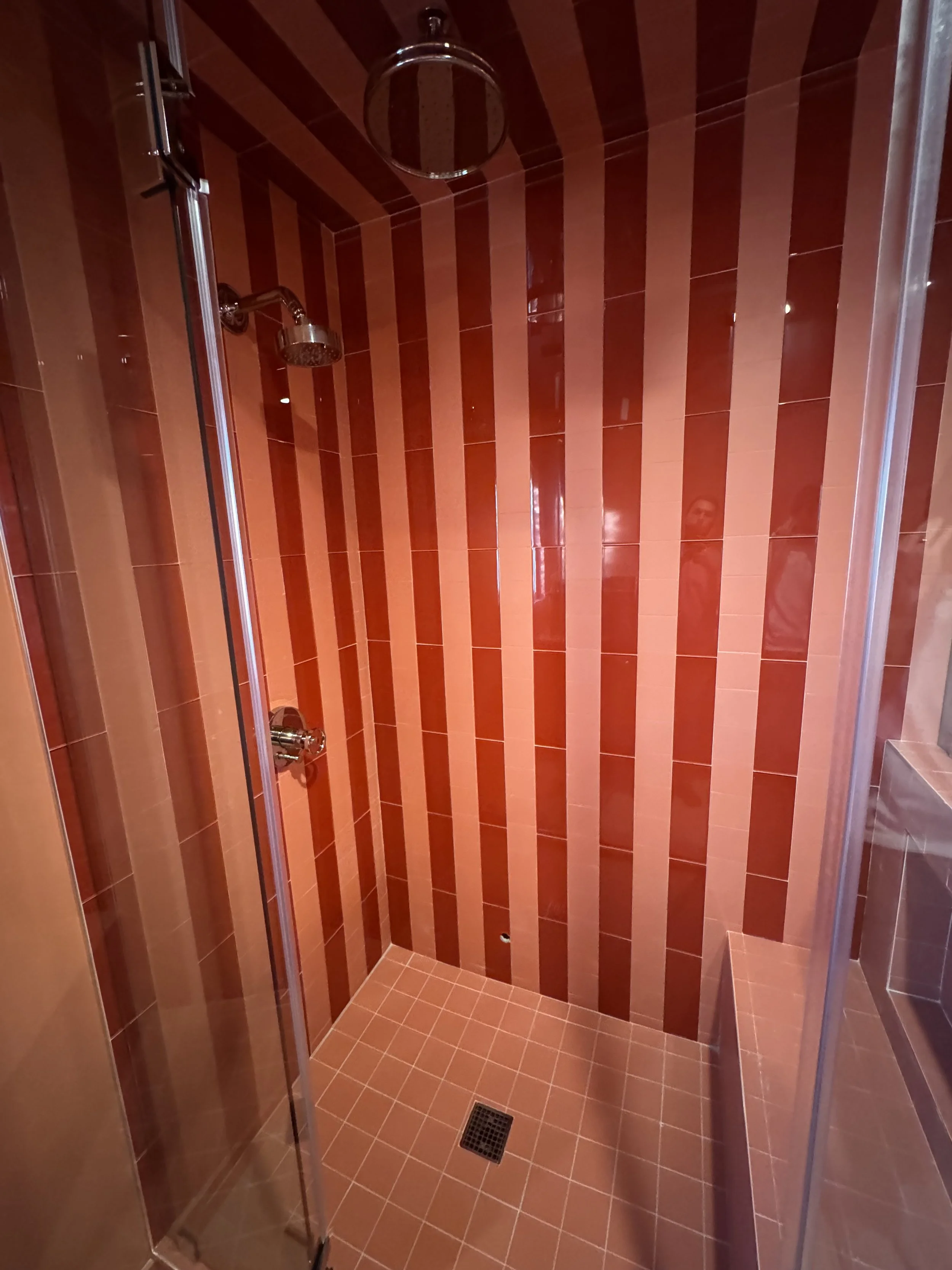 Inside a tiled shower with red and orange vertical stripes, a ceiling-mounted showerhead, and a side bench.