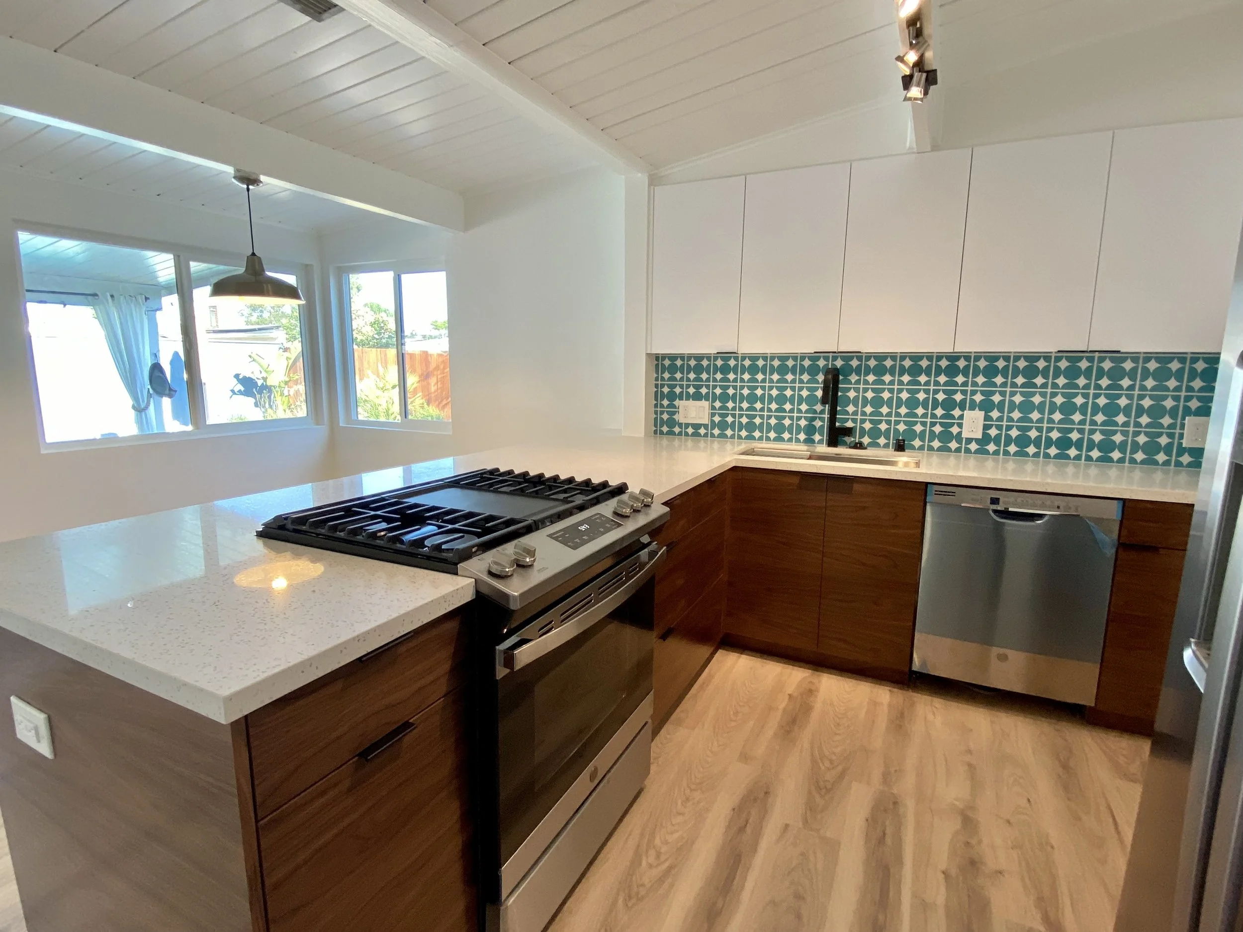 Modern kitchen with wooden cabinets, a speckled white countertop, a stainless steel oven and stove, a small sink with a black faucet, a teal patterned tile backsplash, and a window allowing natural light, with light wood flooring.