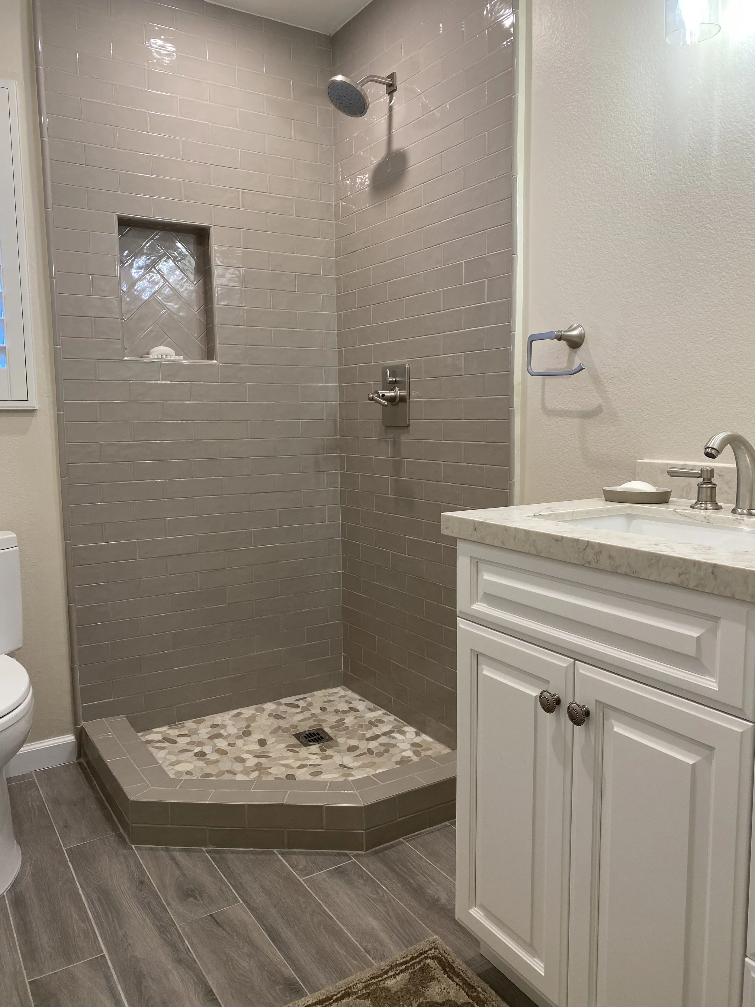 Bathroom with walk-in shower featuring beige tile walls and pebble tile floor, white vanity with marble countertop, and a partial view of toilet.