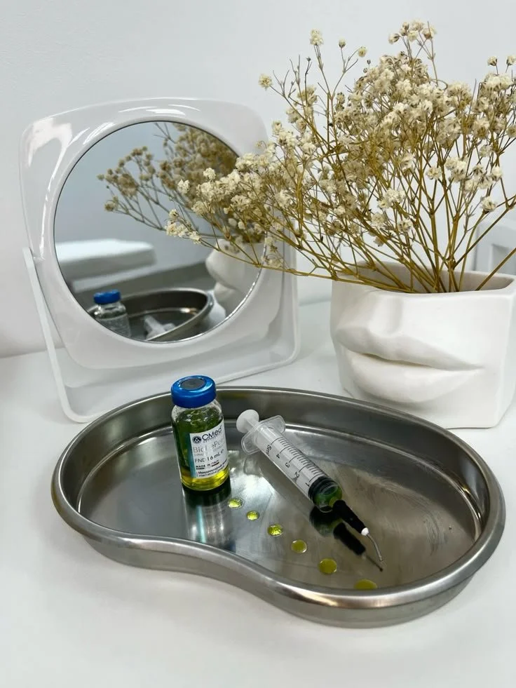 A medical tray with a vial of greenish liquid, a syringe, and yellow pills, beside a white vase with dried white flowers and a mirror reflecting part of the setup.
