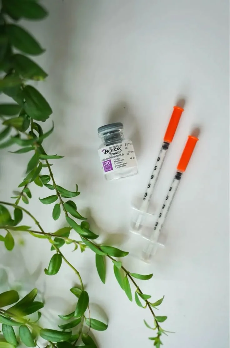 A small jar of Botox cosmetic, two syringes with orange caps, and a leafy green plant on a white surface.
