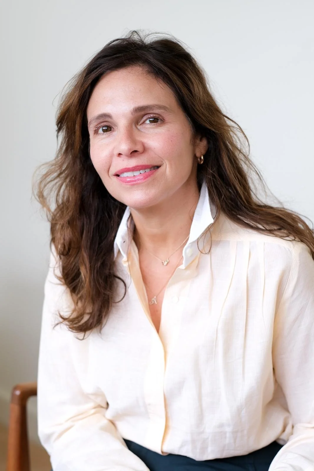 A woman with shoulder-length wavy brown hair, smiling, wearing a white blouse and small gold jewelry, sitting against a plain light wall. Alisa Septimus Boymelgreen LCSW