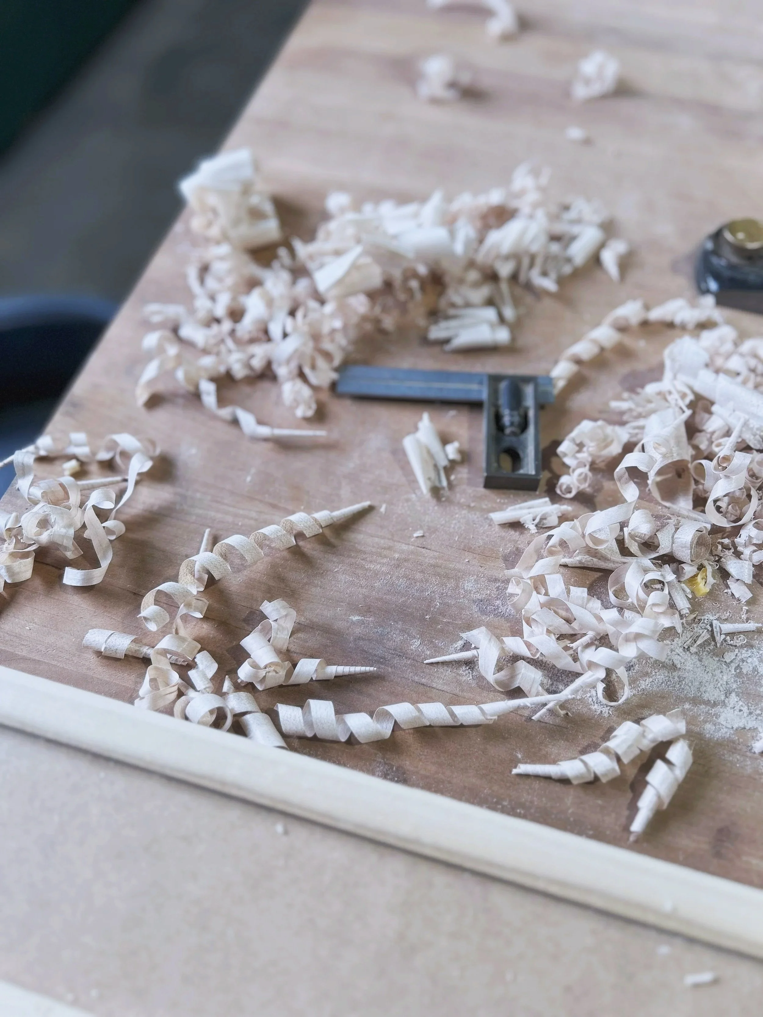 Woodwork shavings and curls scattered on a wooden work surface with a small woodworking plane nearby.