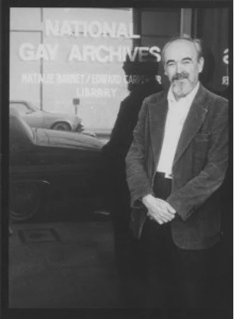 Jim Kepner, a white man with a mustache and goatee, stnads in front of a window that says National Gay Archives.
