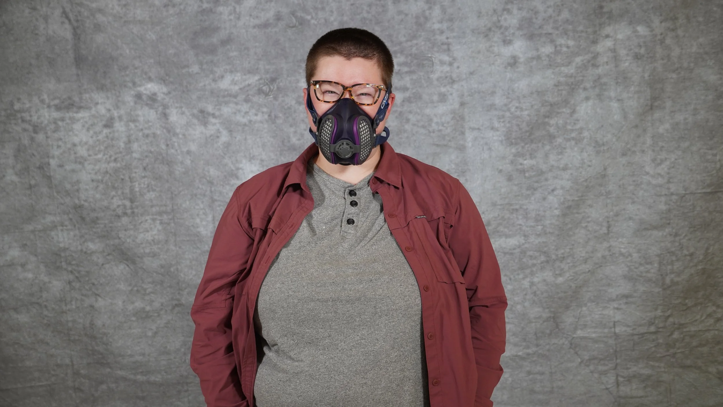 A masc-presenting nonbinary person with short brown hair, tortoiseshell glasses, and a blue and purple P100 mask, wearing a gray shirt and a red button down, stands in front of a gray backdrop.