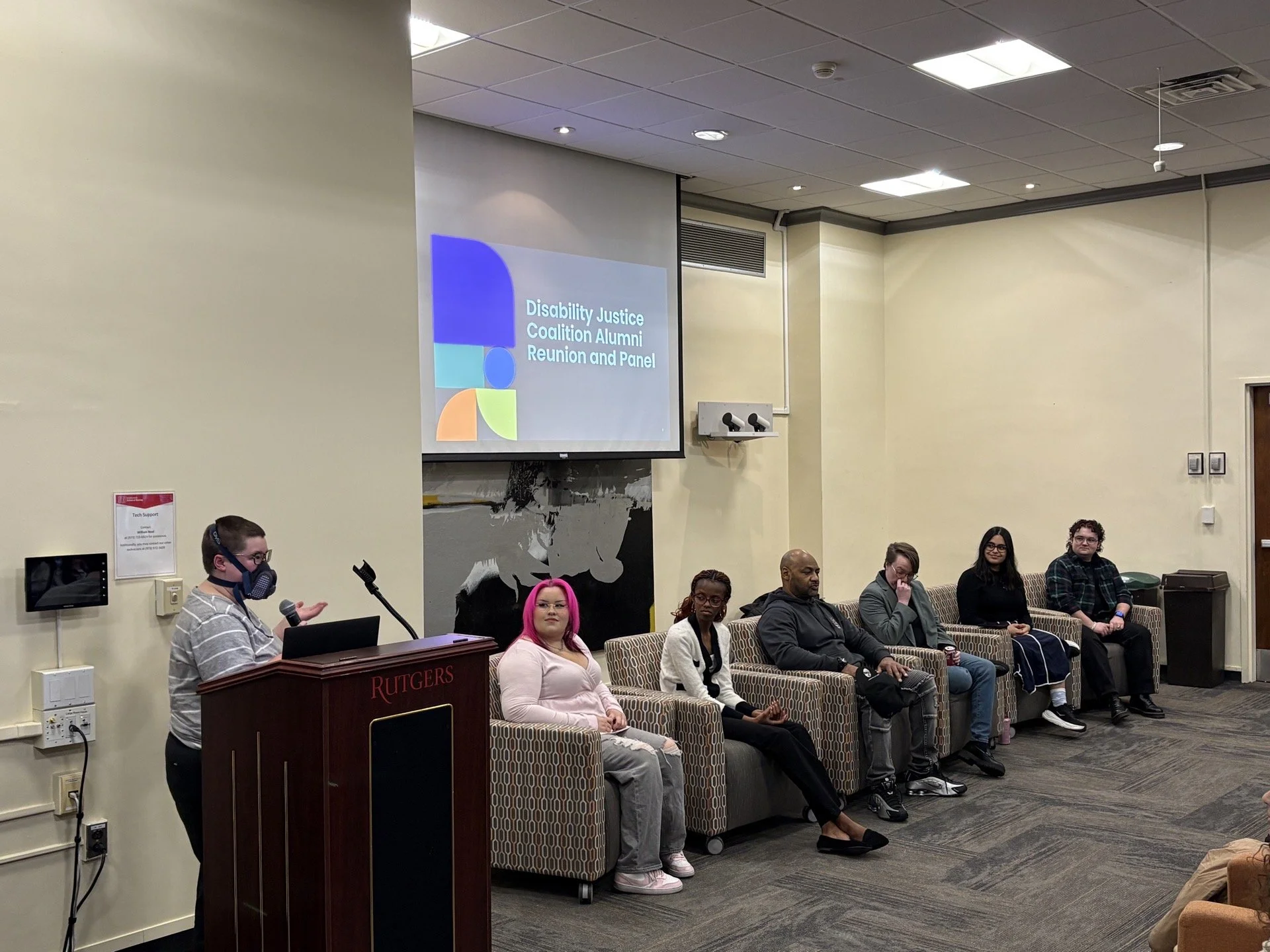 Moira stands at a podium, gesturing to a panel of students sitting in armchairs. A projector screen in the background reads Disability Justice Coalition Alumni Reunion and Panel.