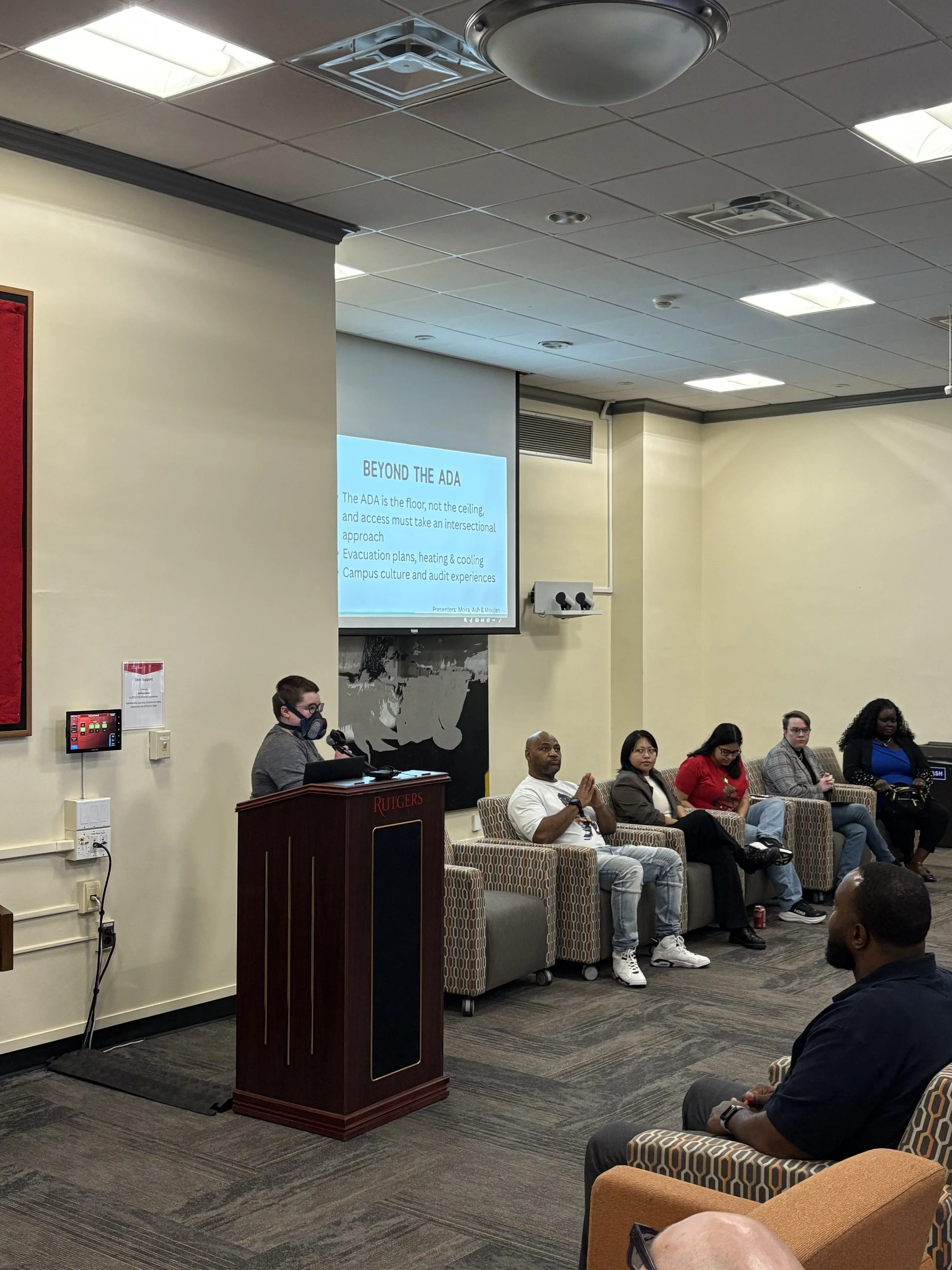 Moira stands at a podium, introducing a slate of student speakers. There is a projected slideshow in the background with the heading "Beyond the ADA."