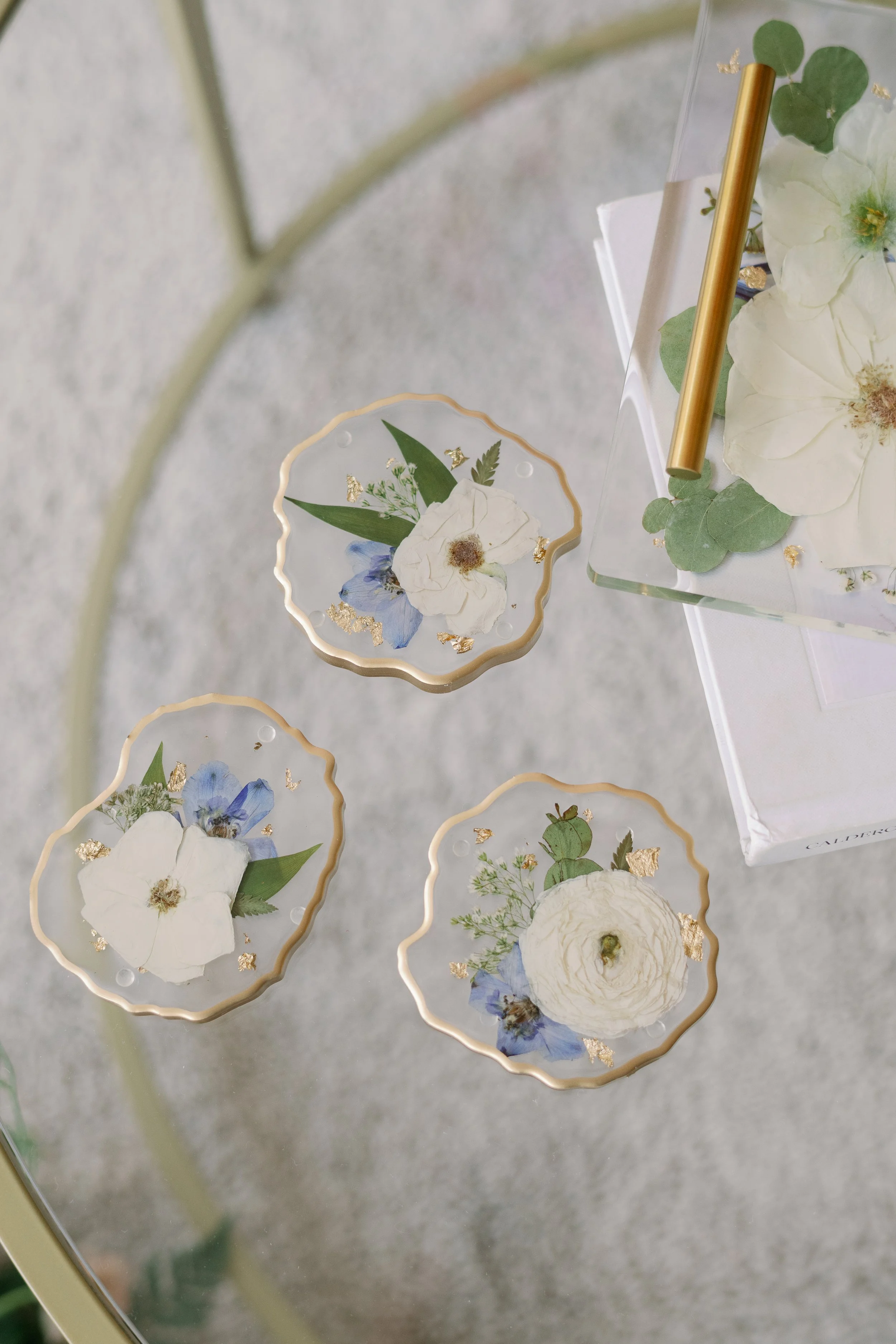 Three gold-rimmed geode resin coasters preserving white wedding flowers, blue petals, greenery, and gold leaf beside a matching floral tray