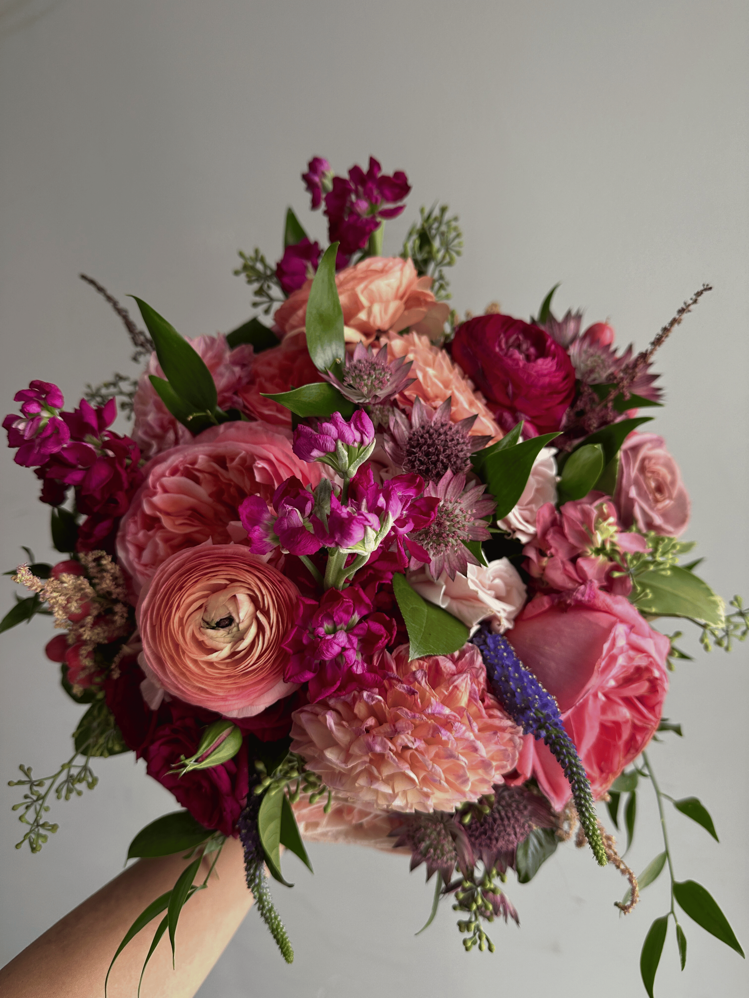 A hand holding a vibrant bouquet of pink, purple, and peach flowers with green leaves against a plain light gray background.