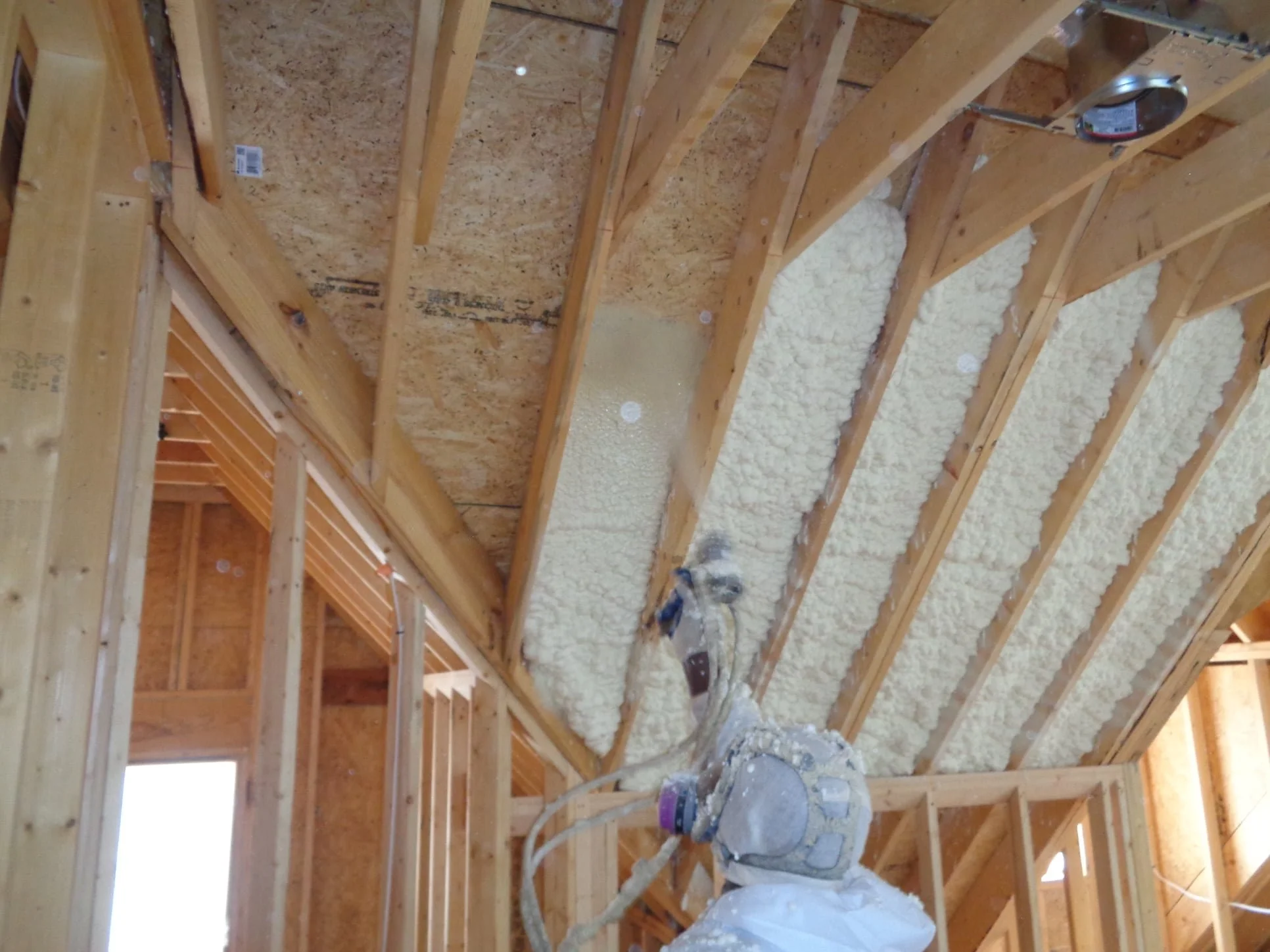 Construction worker installing spray foam insulation between wooden wall studs in a building under construction.