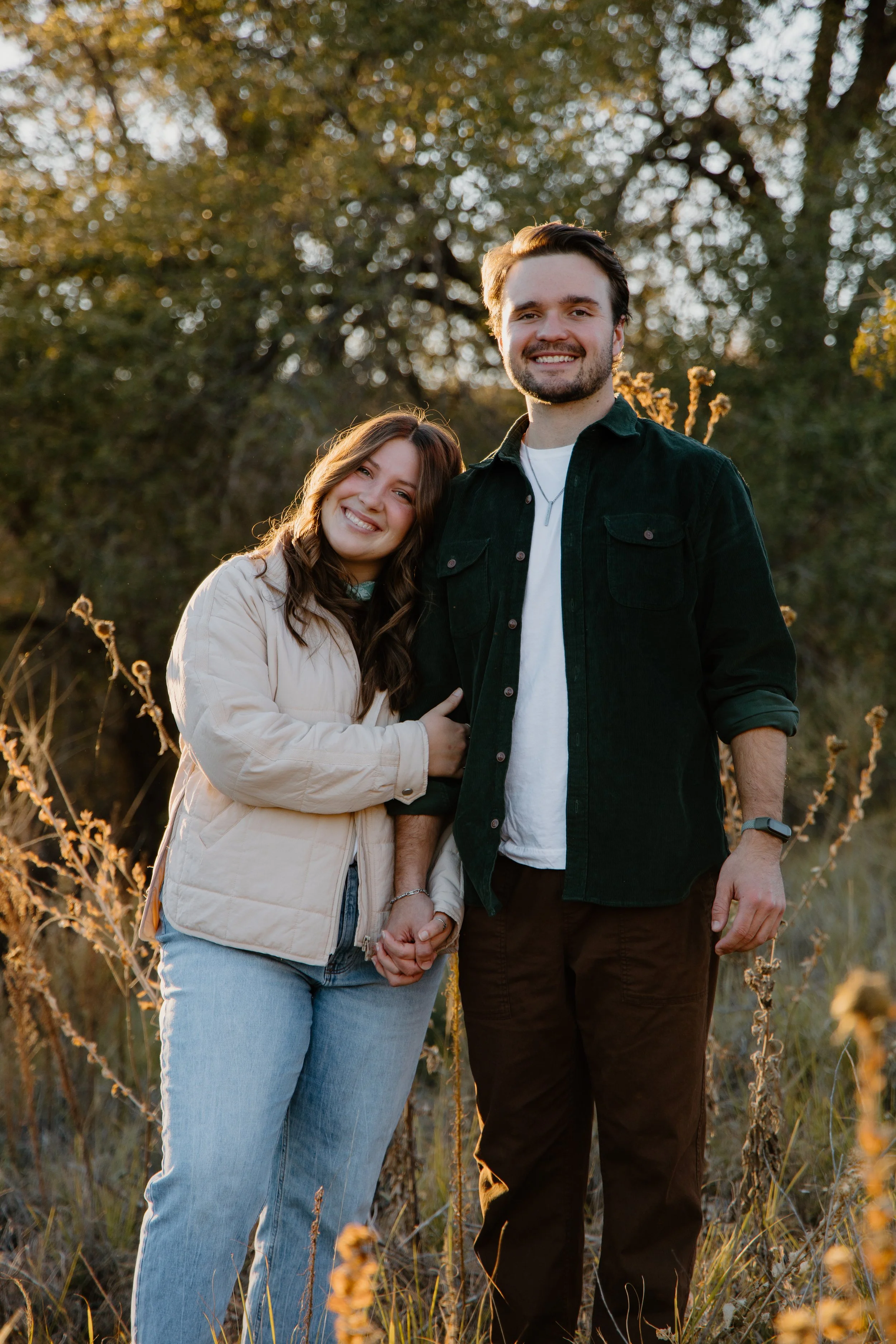 A smiling young woman and a man holding hands outdoors during sunset, with trees and golden light in the background.