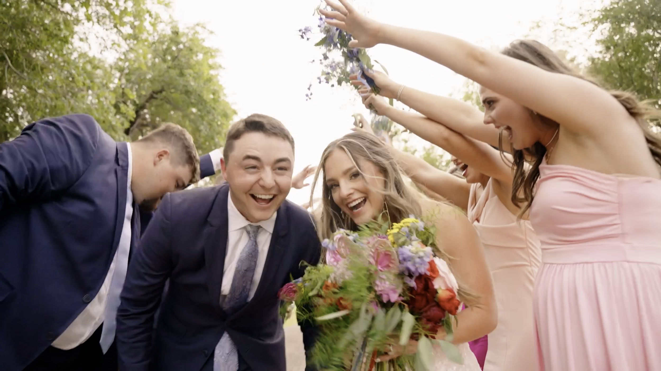 Group of people celebrating outdoors, smiling, surrounding a woman holding a bouquet of flowers, with women raising their hands in the air.