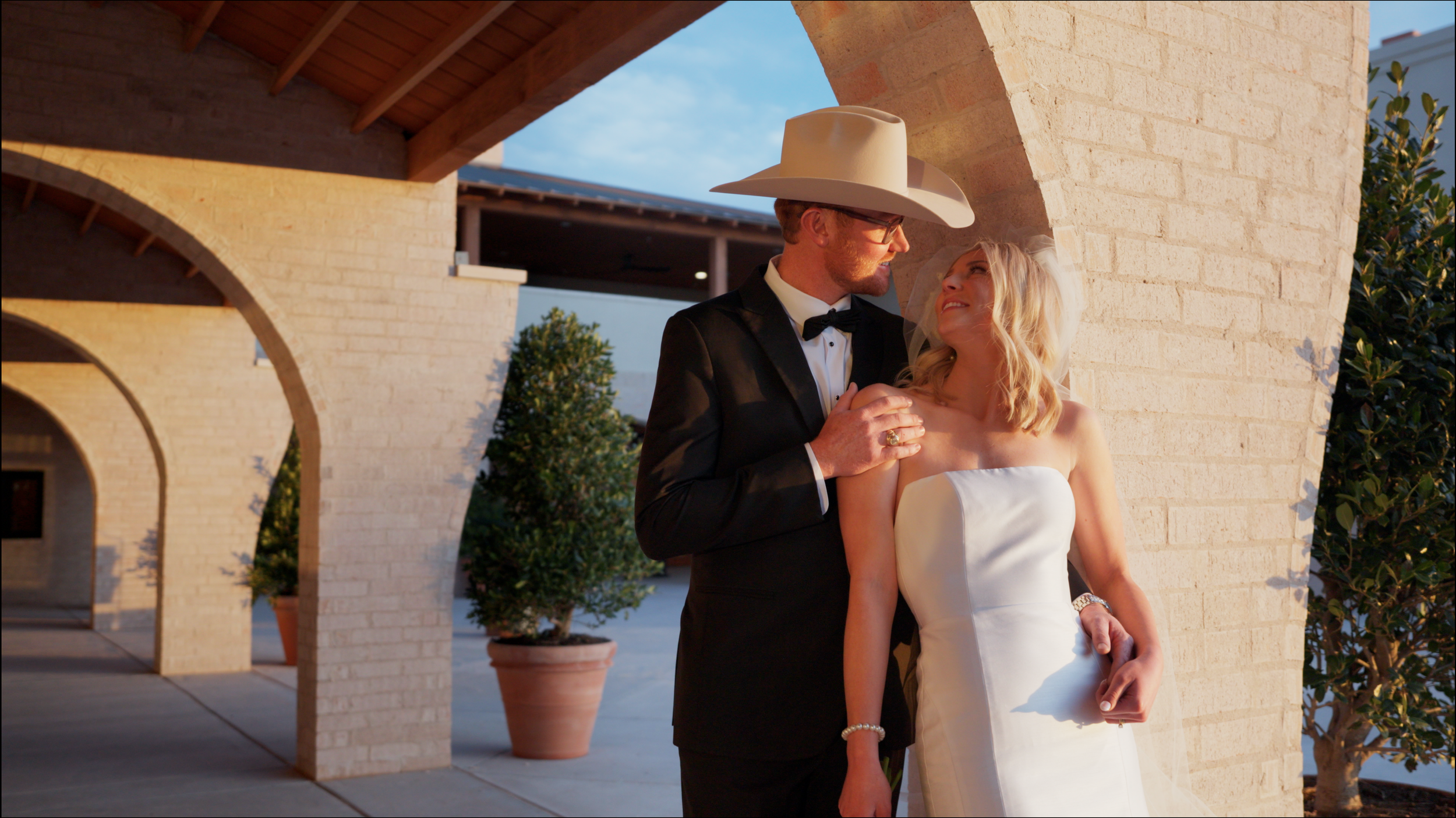 A couple dressed in wedding attire standing close to each other under a brick archway, with the groom wearing a black tuxedo and the bride in a white strapless wedding dress, during sunset.